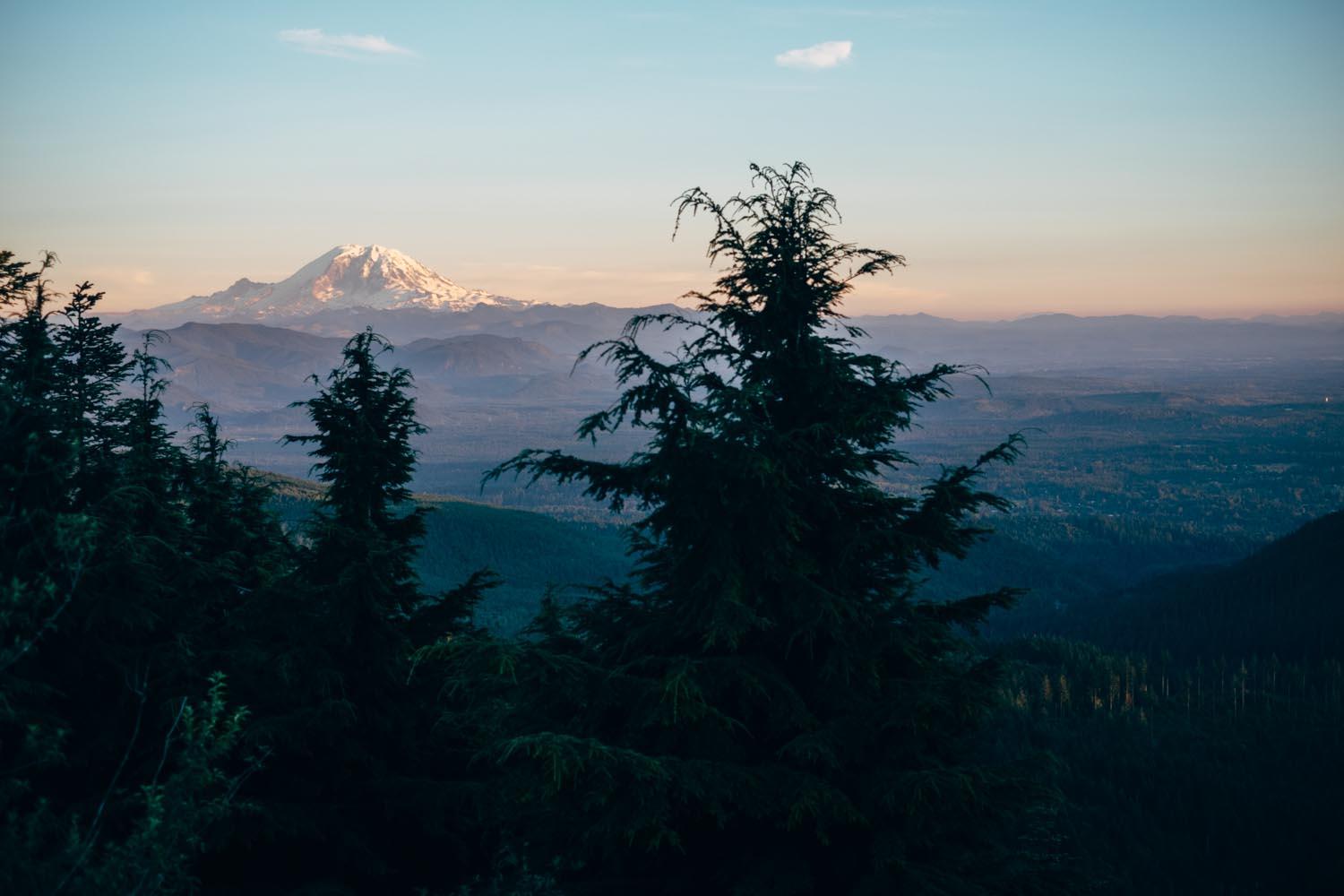 A scenic view of a snow-capped mountain in the distance, surrounded by a vast landscape of evergreen trees and rolling hills under a clear sky during dusk. The foreground features dark green trees framing the view, while the mountain stands prominently against the horizon. Tiger Mountain mountain bike trail.