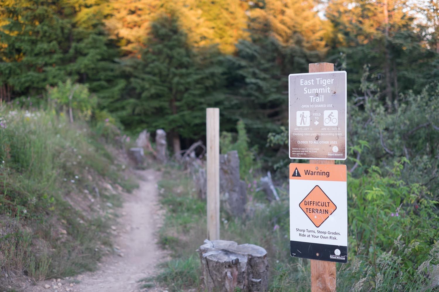 A dirt path leading into a forest, flanked by greenery, with two signs posted. The upper sign indicates the "East Tiger Summit Trail" is open for shared use and provides seasonal information. The lower sign warns of "Difficult Terrain," highlighting sharp turns and steep grades, advising users to ride at their own risk. Tiger Mountain mountain bike trail.