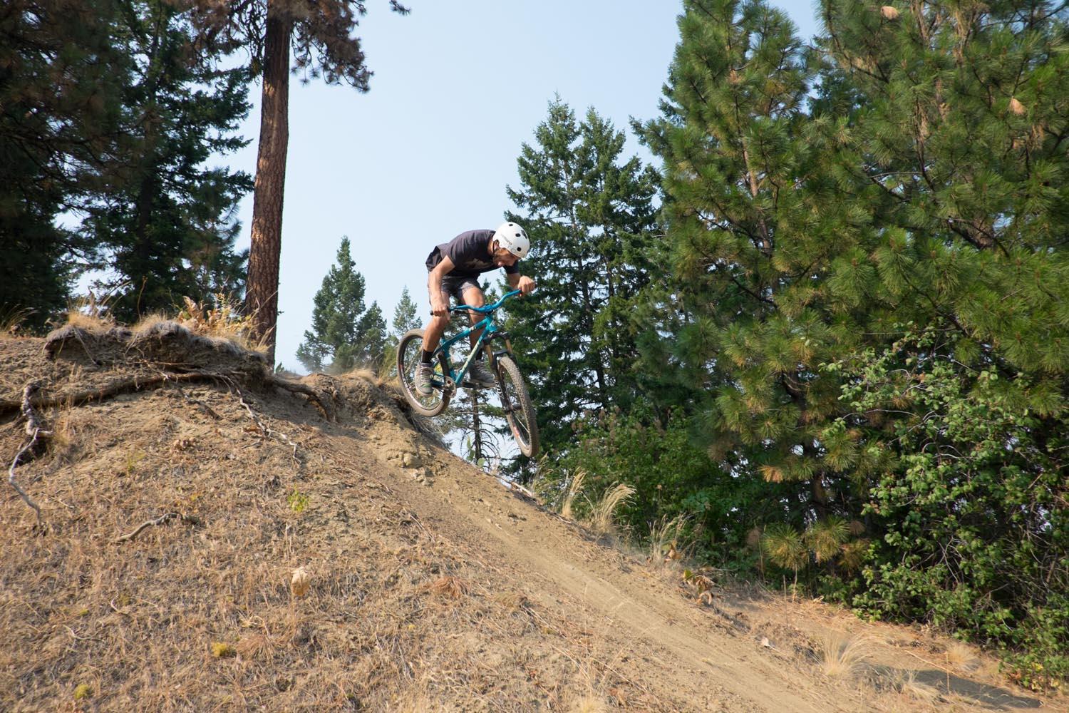 A mountain biker in a helmet jumps off a dirt ramp, with trees in the background. The rider is airborne, showcasing their skills on a rugged, natural trail. Freund Creek mountain bike trail.