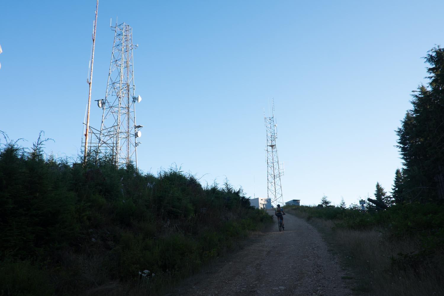 A dirt path leading up a hill with two tall radio towers and a small building at the top. A person on a bike is riding up the path, surrounded by greenery under a clear blue sky. Tiger Mountain mountain bike trail.