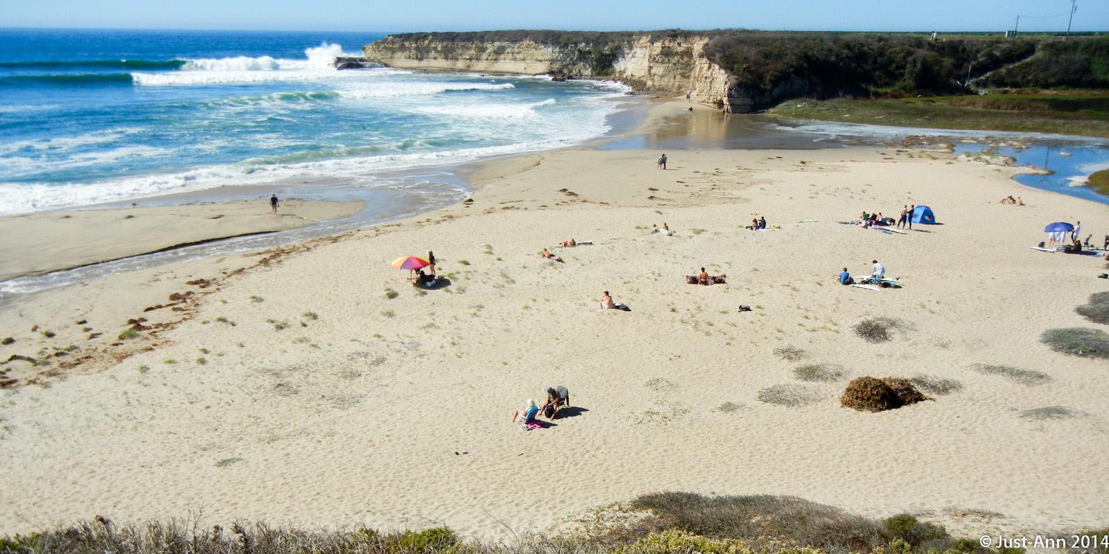 A panoramic view of a sandy beach with gentle waves lapping at the shore. People enjoy the sun and surf, some lounging on towels, while others walk along the water's edge. Colorful umbrellas provide shade, and rocky cliffs can be seen in the background under a clear blue sky. Wilder Ranch State Park mountain bike trail.