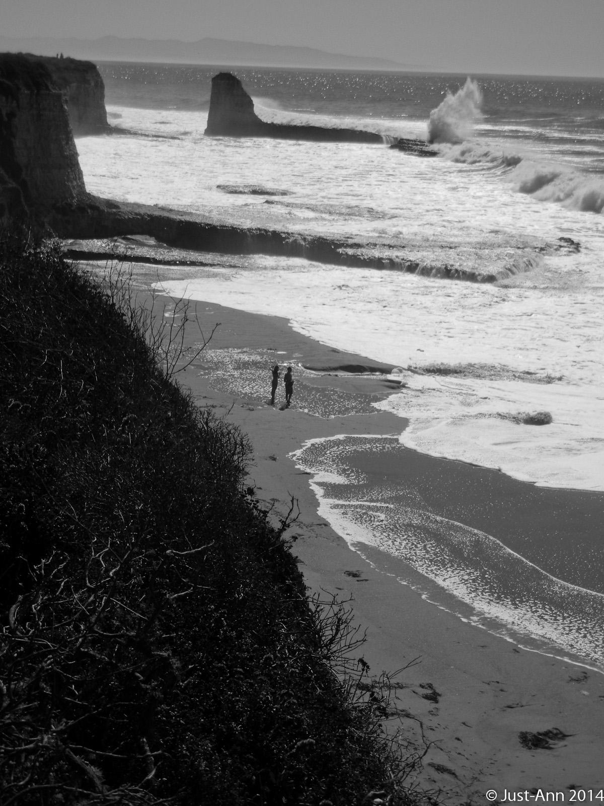 A black and white image of a sandy beach with two people walking along the shore. Towering rock formations rise from the ocean in the background, with waves crashing against them. The scene captures a peaceful coastal landscape under a bright sky. Wilder Ranch State Park mountain bike trail.