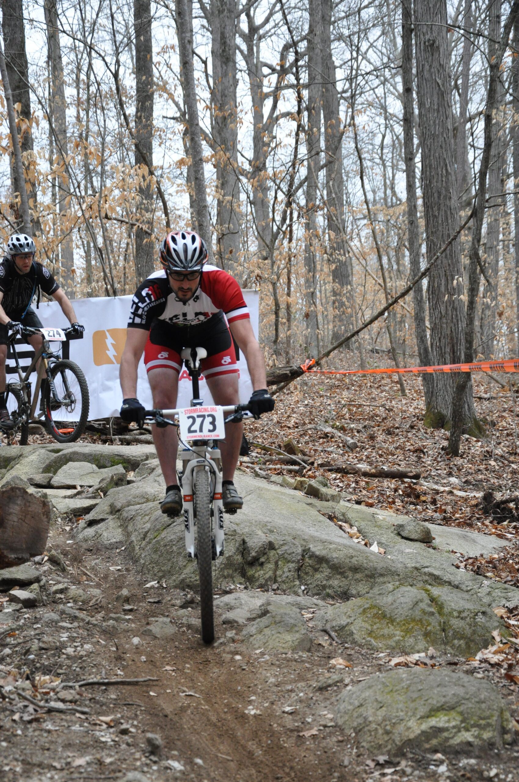A mountain biker navigating rocky terrain during a race, with a second competitor in the background. The wooded area features bare trees and scattered leaves, creating a natural setting for the event. The biker wears a red and black cycling jersey and a helmet, focused on maintaining balance over the rocks. A banner and orange tape indicate the race course. San-lee Park mountain bike trail.