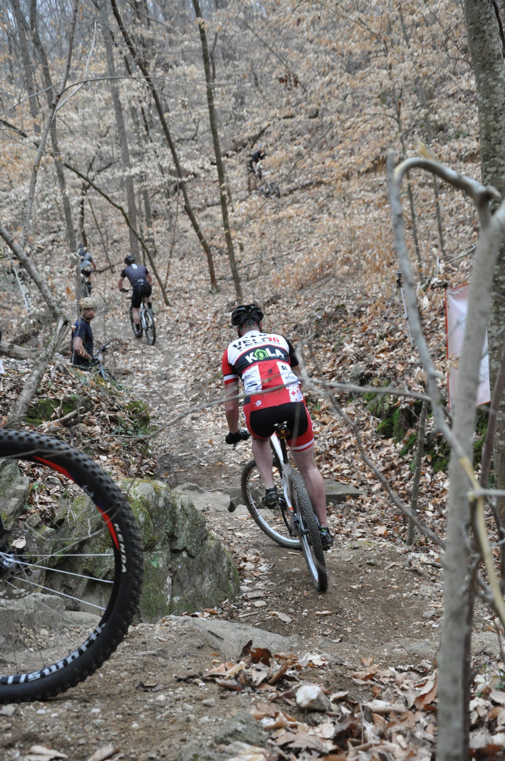 A mountain biker navigates a rocky trail in a forested area during a race, surrounded by trees with few leaves and scattered fallen leaves on the ground. Other cyclists are visible in the background. San-lee Park mountain bike trail.