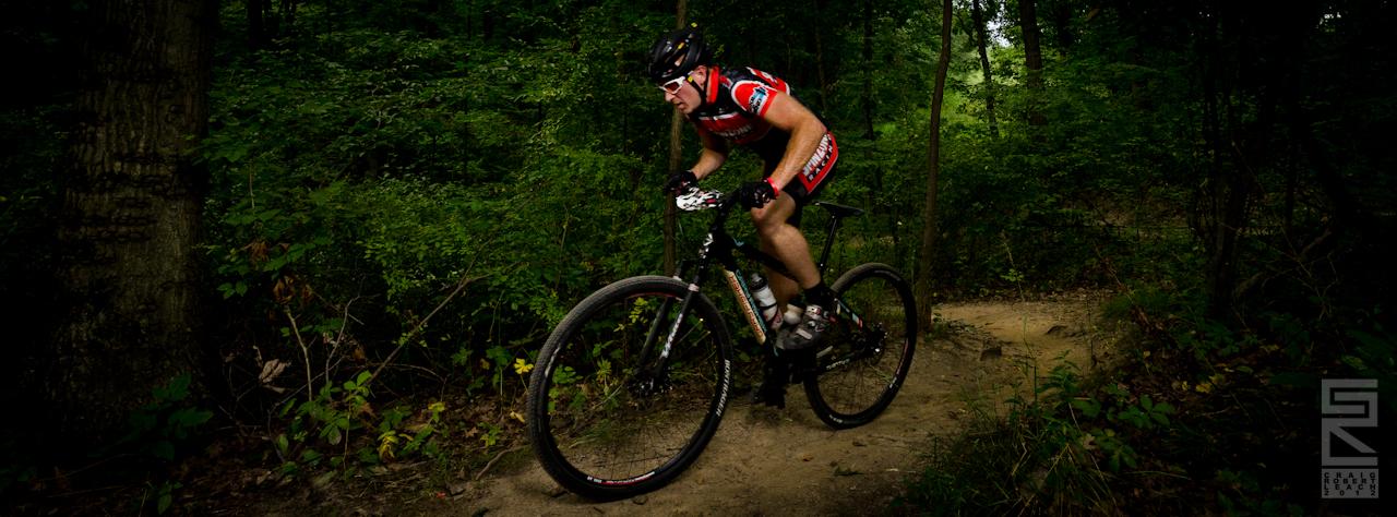 A mountain biker navigating a winding dirt trail through a lush green forest, wearing a red and black cycling outfit and a helmet, demonstrating speed and agility.