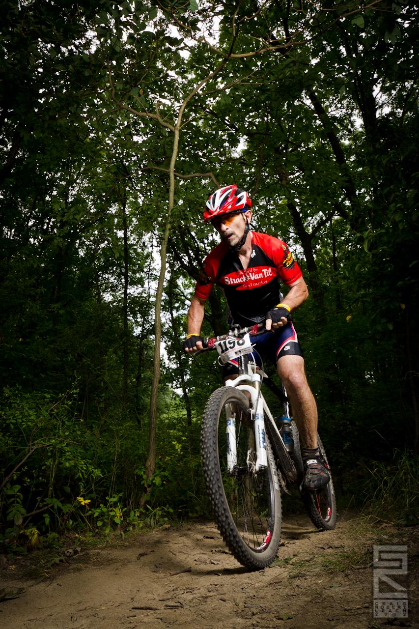 A male mountain biker in a red and black jersey navigates a dirt trail through a wooded area. He wears a helmet and is focused on the path ahead, with trees surrounding him. The bike features thick tires suitable for off-road riding.