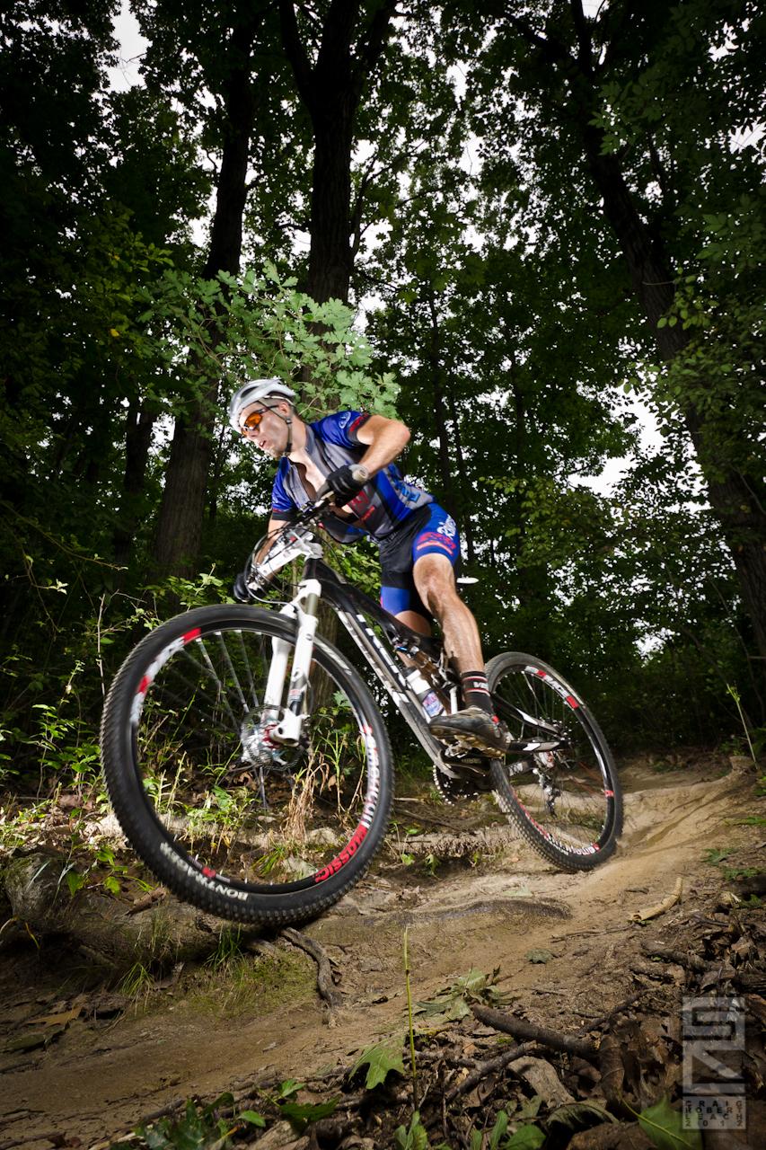 A cyclist in a blue jersey and helmet rides a mountain bike on a dirt trail, surrounded by greenery and trees. The cyclist is captured in motion, leaning forward as they navigate the terrain, with a slight spray of dirt rising from the back wheel.