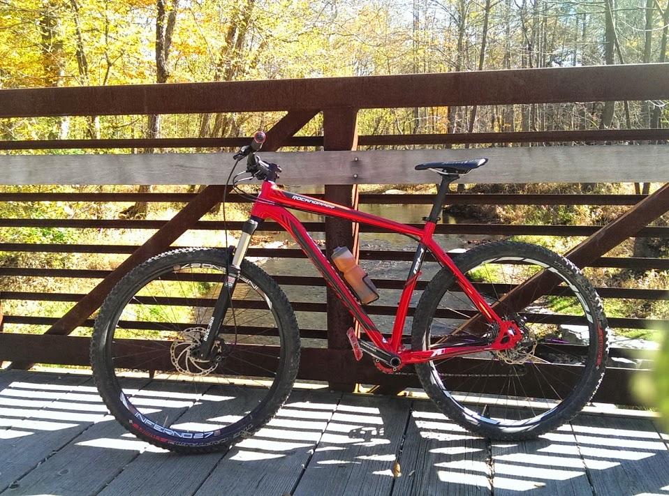 Specialized Rockhopper: A red mountain bike is parked on a wooden bridge, surrounded by trees with autumn foliage. The bike features thick tires and a water bottle mounted on the frame. In the background, a gently flowing stream can be seen through the slatted design of the bridge.