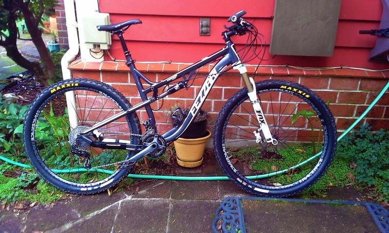 Fezzari Cascade Peak: A mountain bike with a black and silver frame, equipped with Maxxis tires, is leaning against a red wall. The bike features front suspension and a gear system, and is positioned on a paved outdoor area with some greenery and a potted plant in the background.
