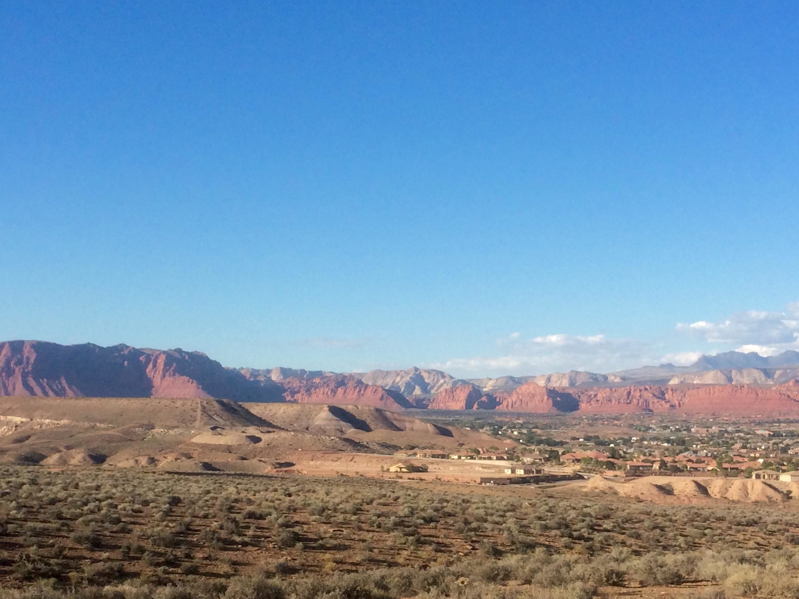 A panoramic view of a desert landscape featuring red mountains in the background under a clear blue sky. The foreground includes sparse vegetation and a small town nestled among the hills. Zen Trail mountain bike trail.