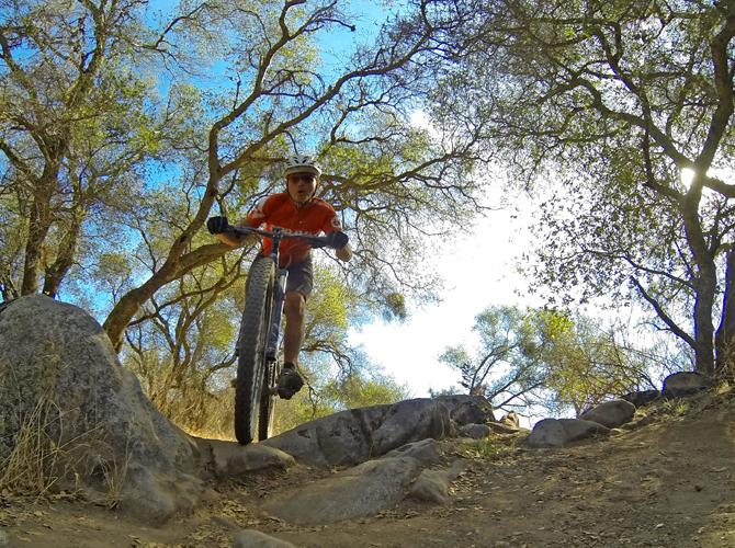 A person riding a mountain bike over rocky terrain surrounded by trees, with sunlight filtering through the foliage. The cyclist is mid-jump, demonstrating skill and agility. Granite Bay Trail mountain bike trail.