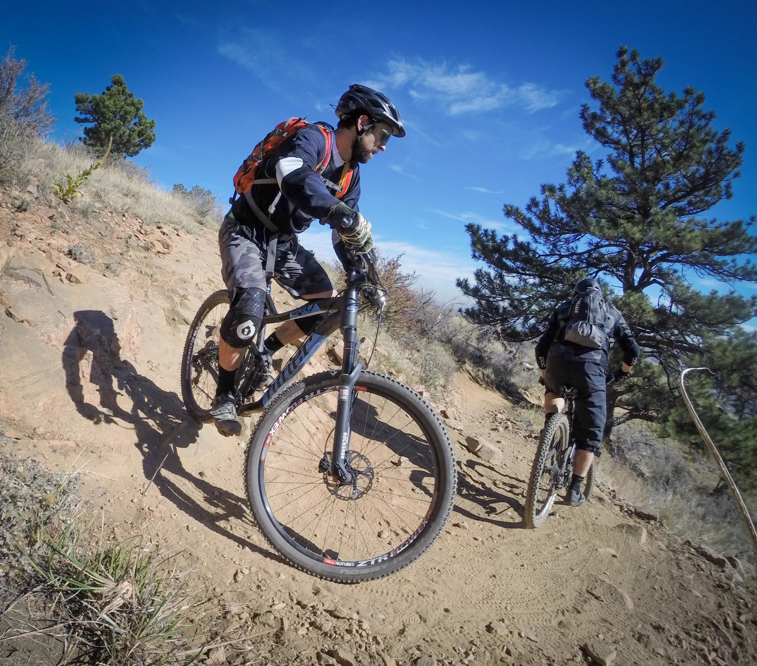 Two mountain bikers navigate a rocky trail on a sunny day. One rider is in the foreground, wearing a helmet and knee pads, focusing on the path ahead, while the second rider is slightly behind, moving up the incline. Pine trees and a clear blue sky are visible in the background, emphasizing the outdoor adventure setting. Apex Park mountain bike trail.