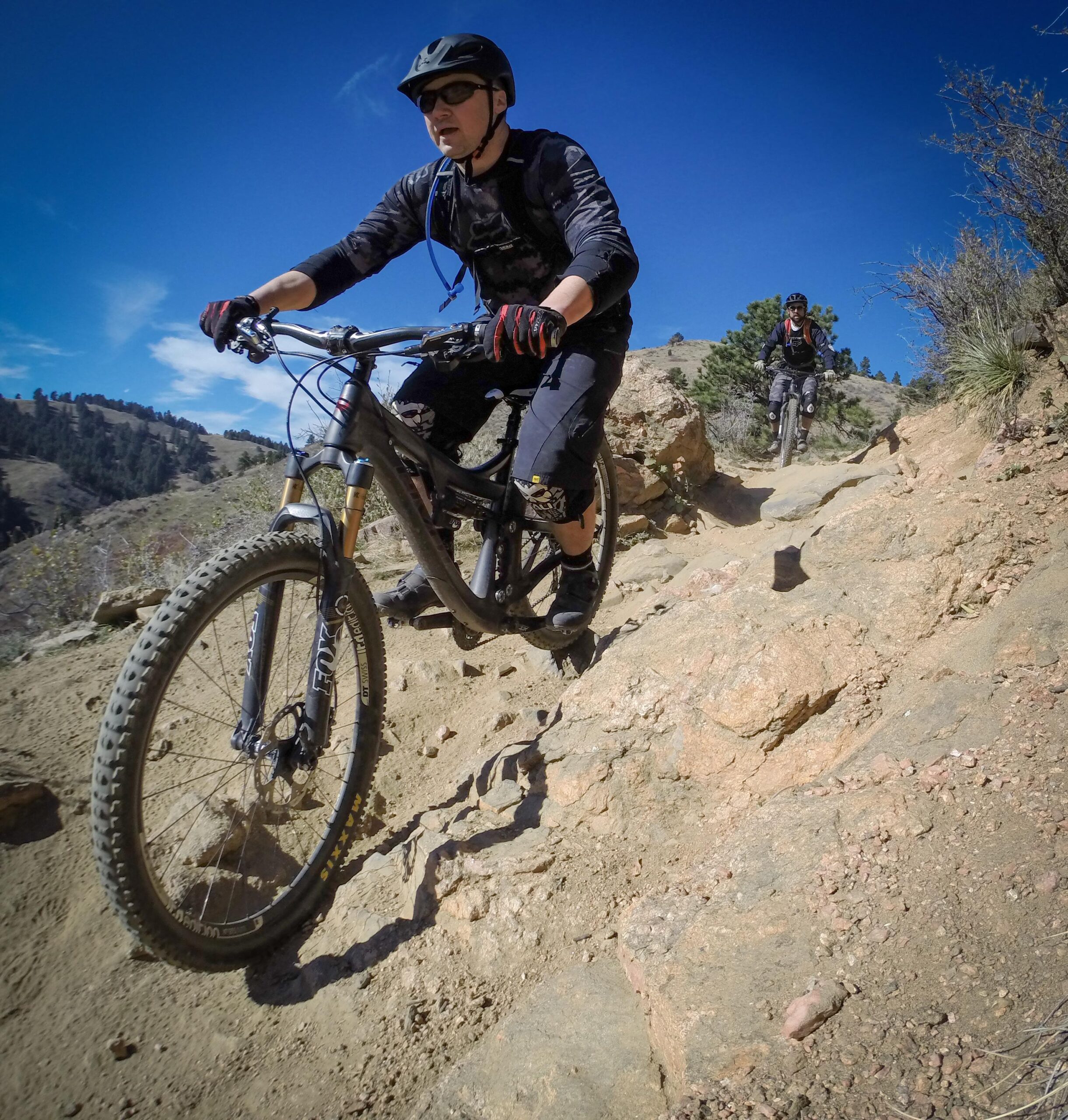 A mountain biker navigating a rocky trail on a sunny day, wearing a helmet and protective gear. Another rider can be seen in the background. The landscape features hills and trees under a blue sky. Apex Park mountain bike trail.