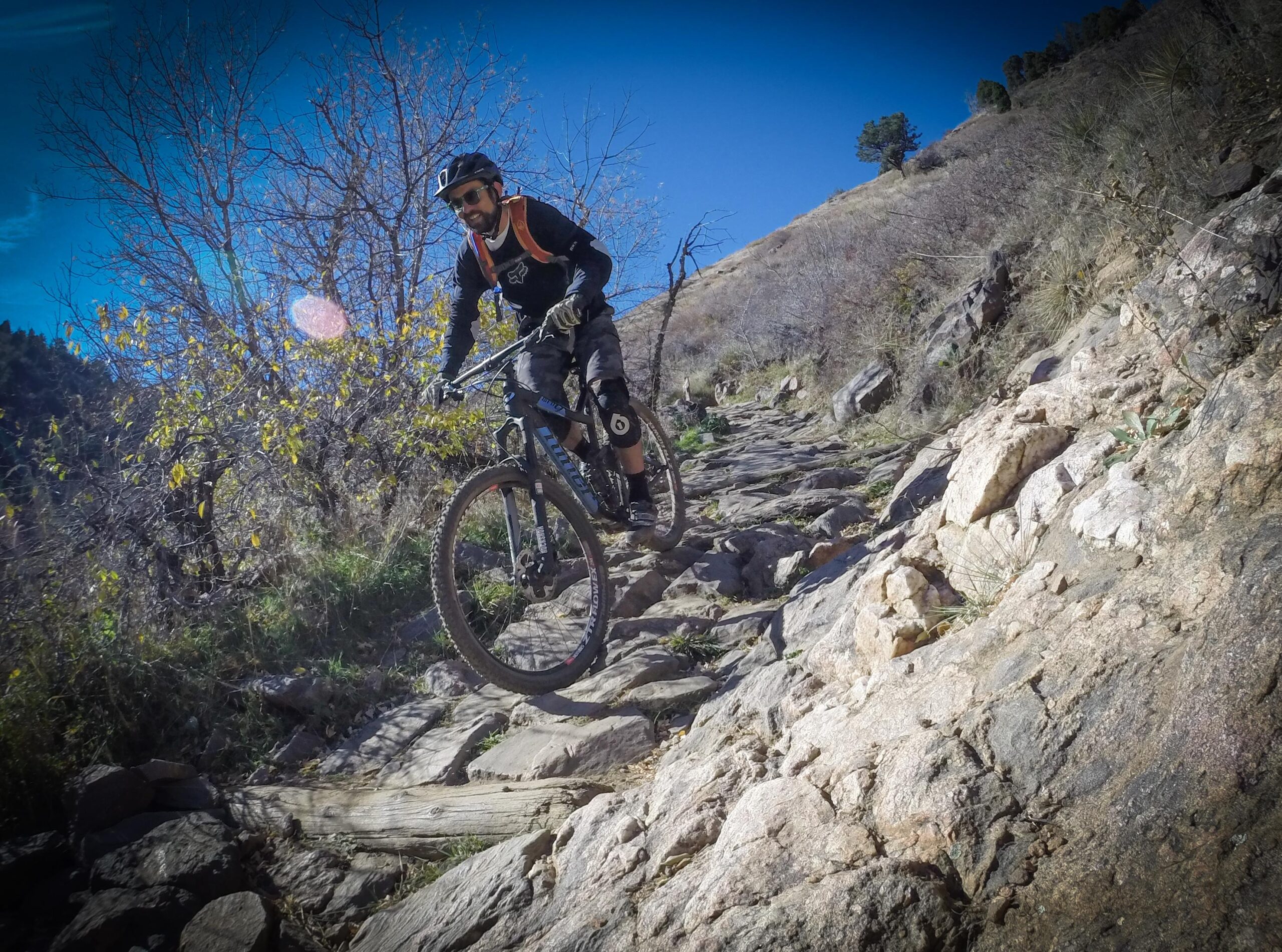 A mountain biker navigating a rocky trail with dirt and grass surrounding the path. The cyclist wears a helmet and protective gear, and the background features a clear blue sky with sparse trees. Apex Park mountain bike trail.