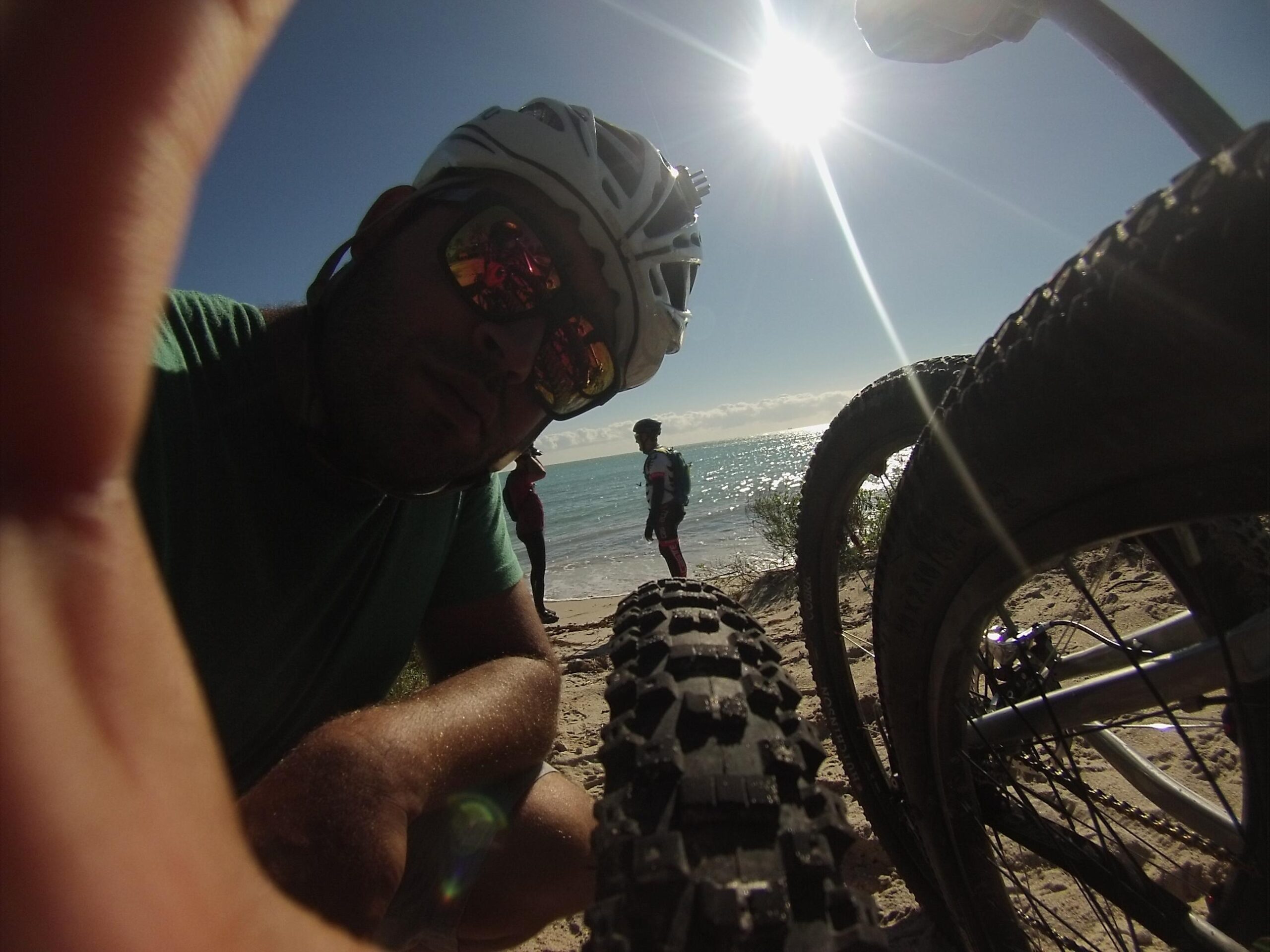 A close-up selfie of a person wearing a bicycle helmet and sunglasses, with a bike tire in the foreground. In the background, another person stands by the water's edge with the sun shining brightly overhead, creating a vibrant outdoor scene. The beach setting features sandy shores and distant waves. Virginia Key North Point mountain bike trail.