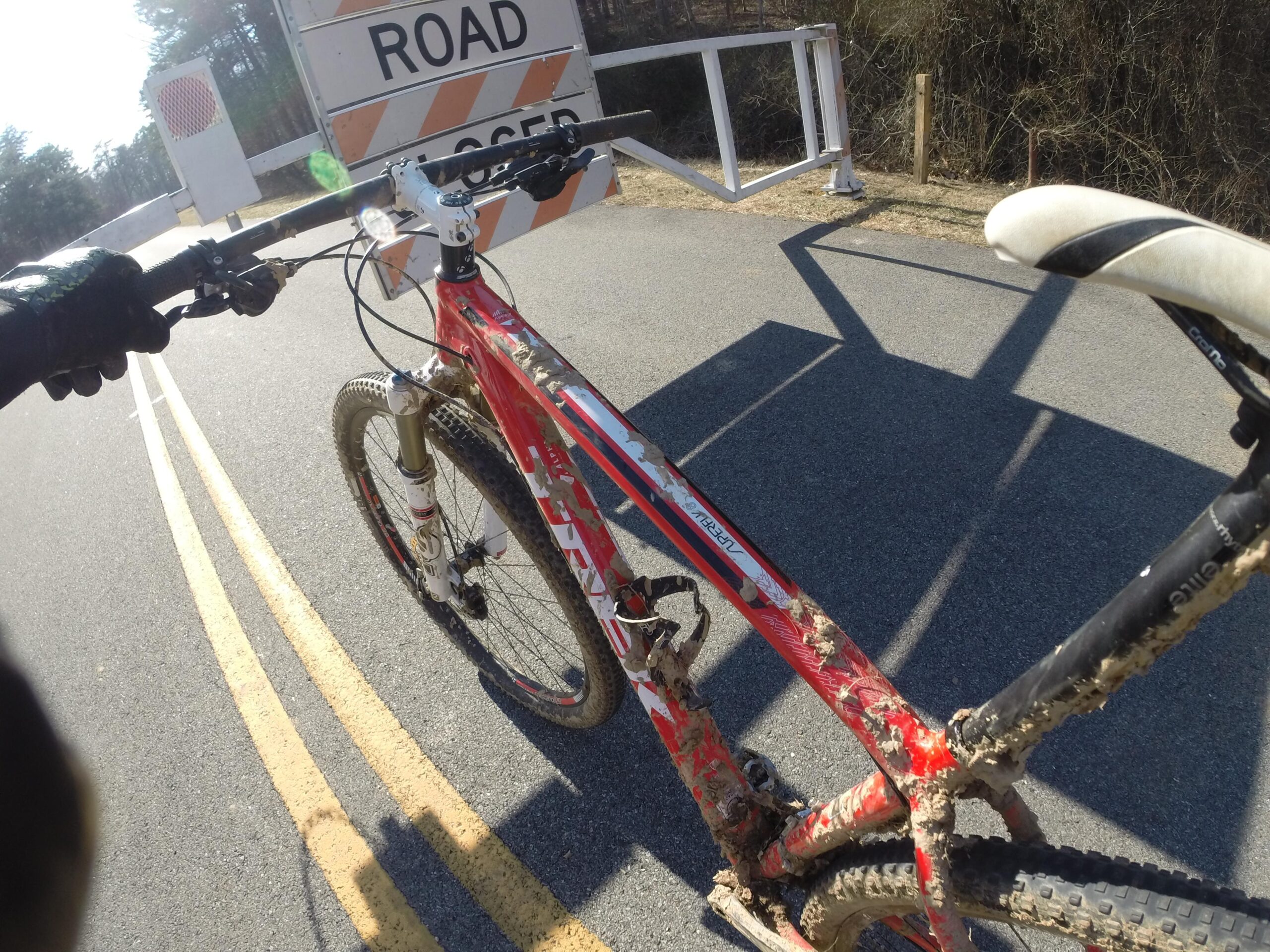 Trek Superfly 8: A close-up view of a mountain bike with a red frame, positioned on a road with a "Road Closed" sign in the background. The bicycle shows signs of dirt and mud, indicating recent off-road use. The focus is on the handlebars and front wheel of the bike, with yellow lane markings visible on the asphalt.