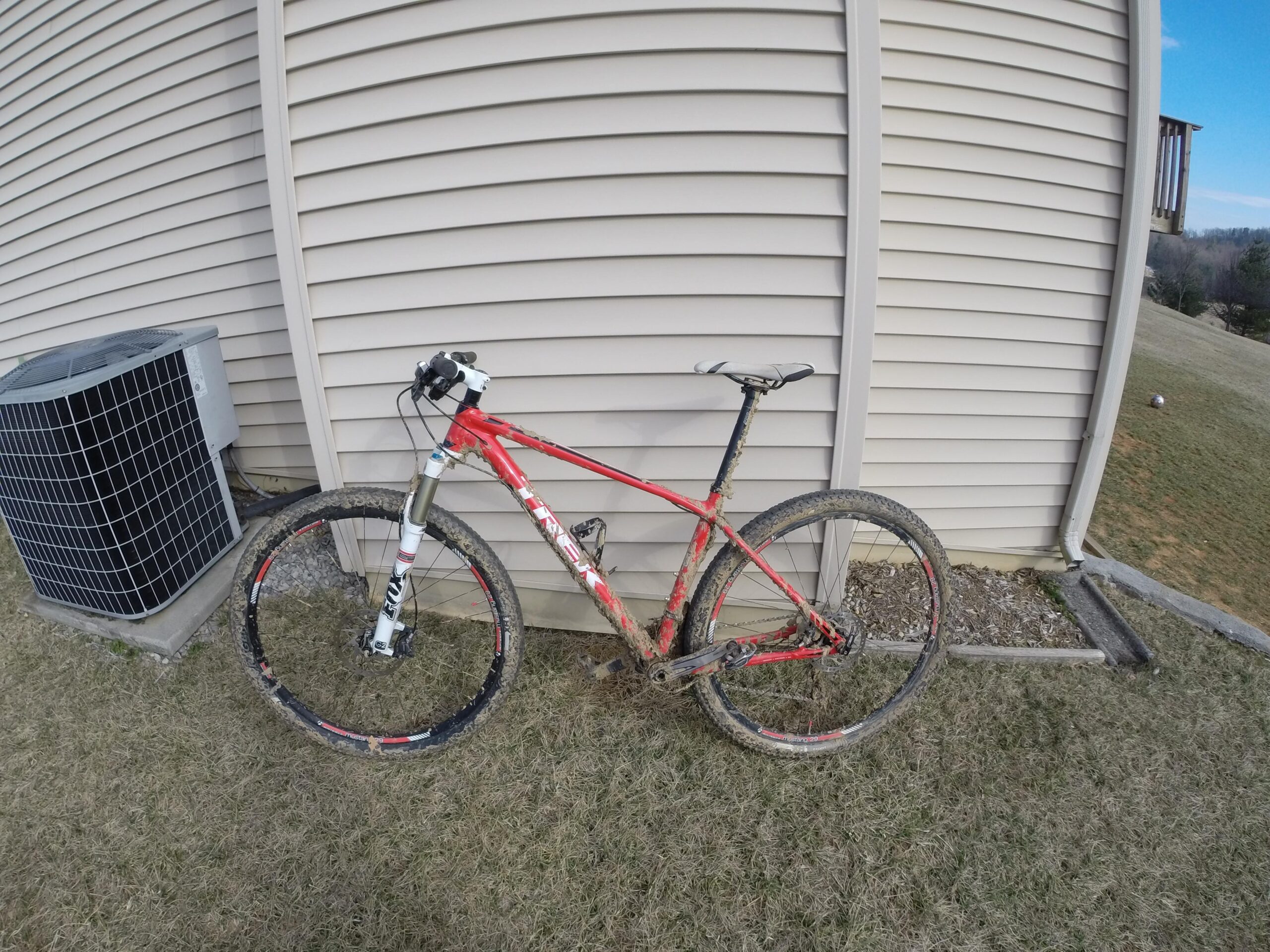 Trek Superfly 8: A red mountain bike leaning against a beige siding wall, next to an air conditioning unit. The bike is covered in mud, indicating recent use on rough terrain. In the background, a grassy area extends into the distance with a clear blue sky.