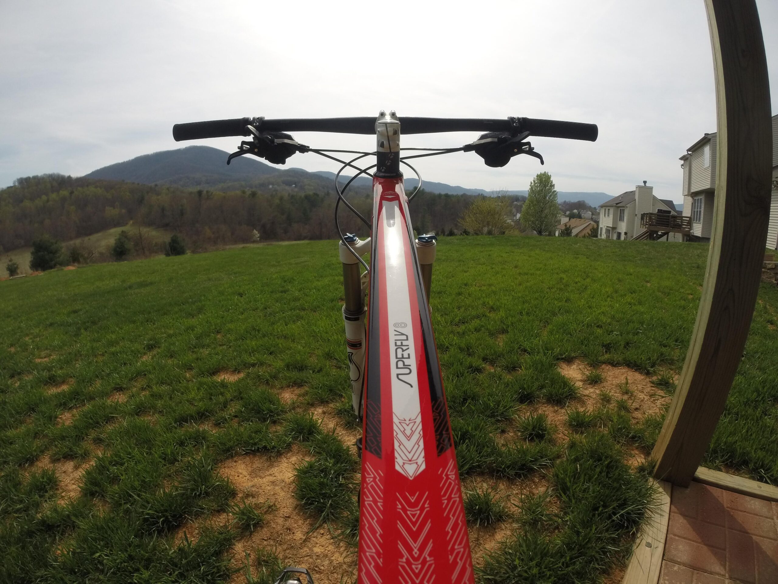 Trek Superfly 8: A mountain bike positioned on green grass, with a view of rolling hills and distant mountains in the background. The handlebars and part of the bike frame are prominent in the foreground, showcasing a vibrant red and white design. The scene is set on a sunny day, with a few homes visible in the distance.