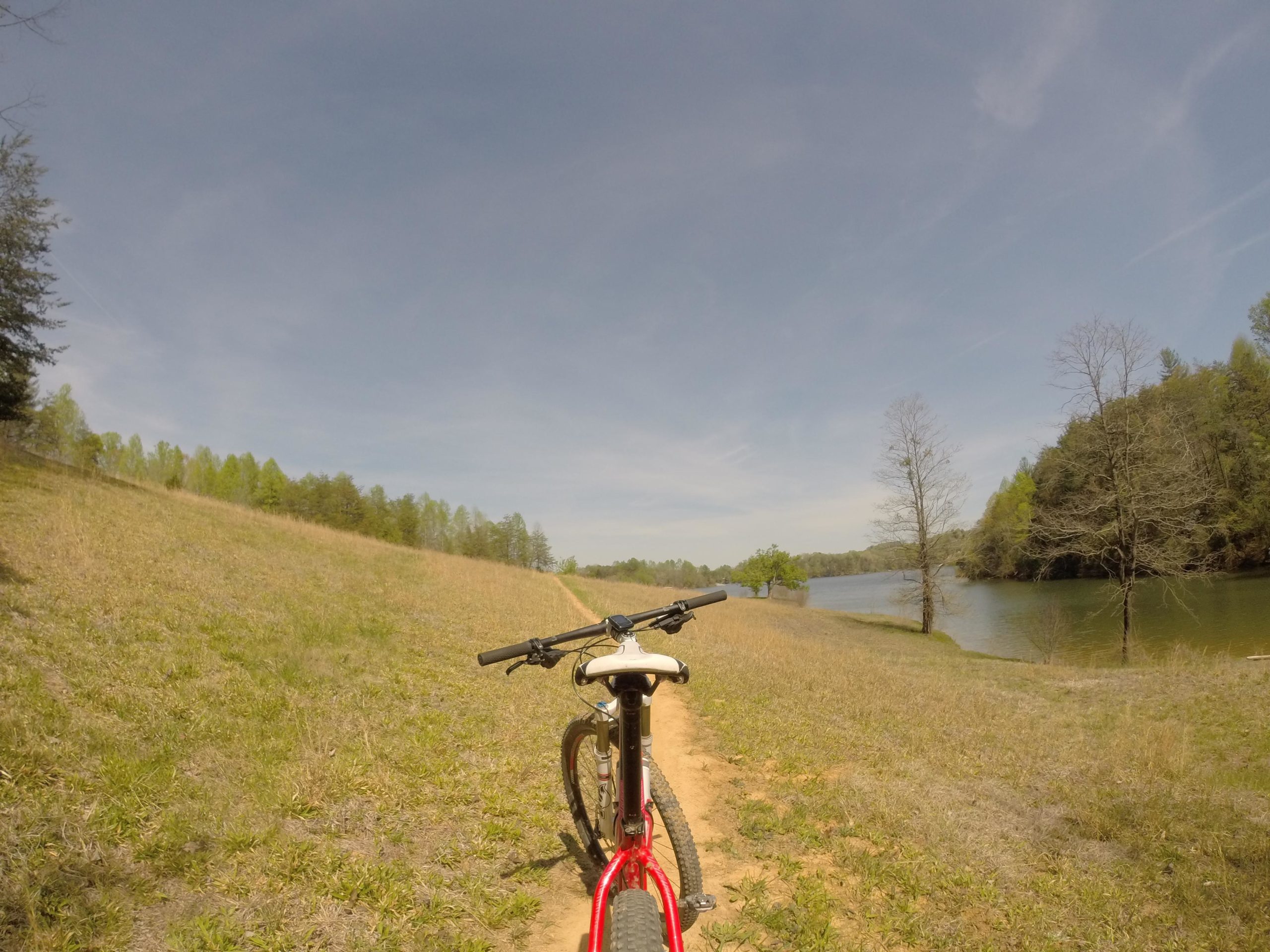 A mountain bike positioned on a dirt path next to a calm lake, with lush greenery and trees in the background under a clear blue sky. Warrior Creek mountain bike trail.