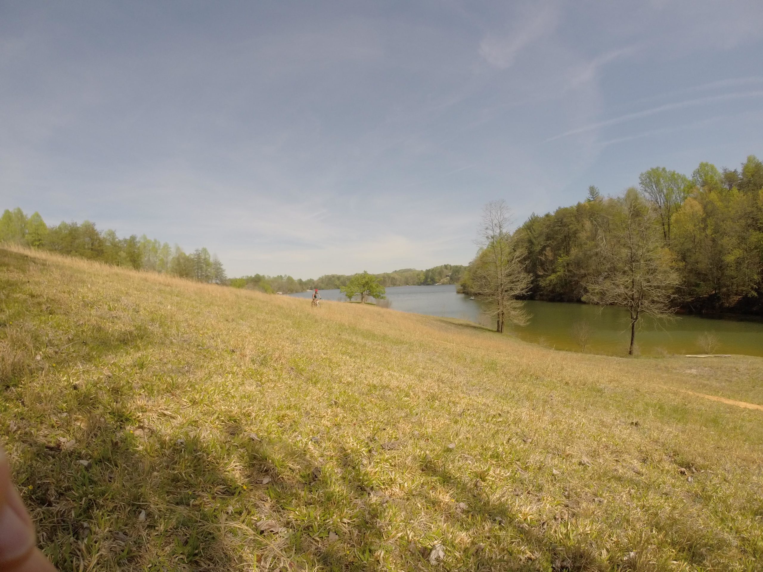 A scenic view of a grassy hillside sloping down to a calm lake, with a few trees on the shore and lush greenery in the background. A person can be seen biking in the distance. The sky is clear with soft clouds, suggesting a pleasant day. Warrior Creek mountain bike trail.