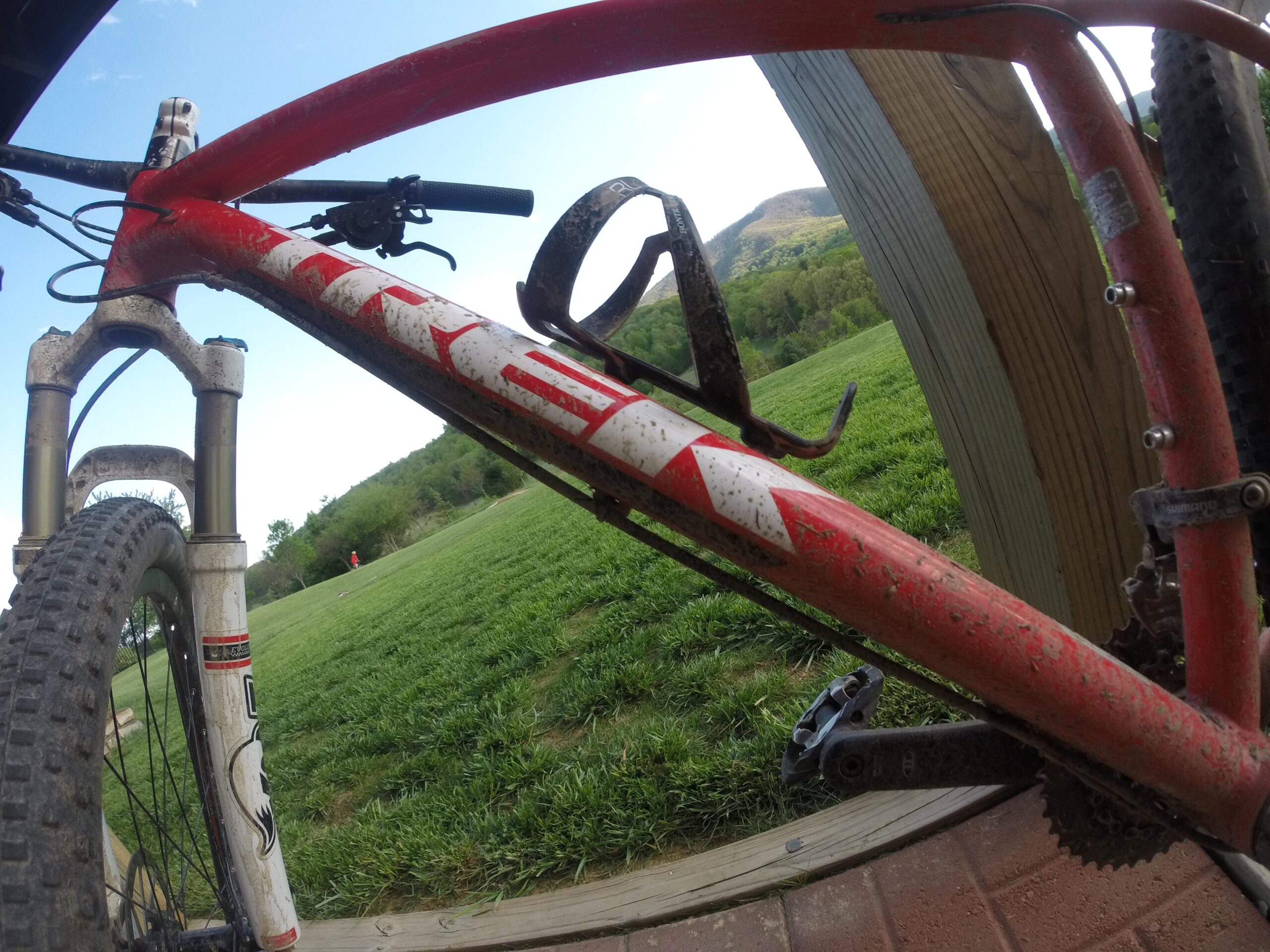 Trek Superfly 8: A close-up view of a red mountain bike with a dirty frame and tires, positioned against a wooden structure. The background features a grassy field and distant hills under a clear blue sky.