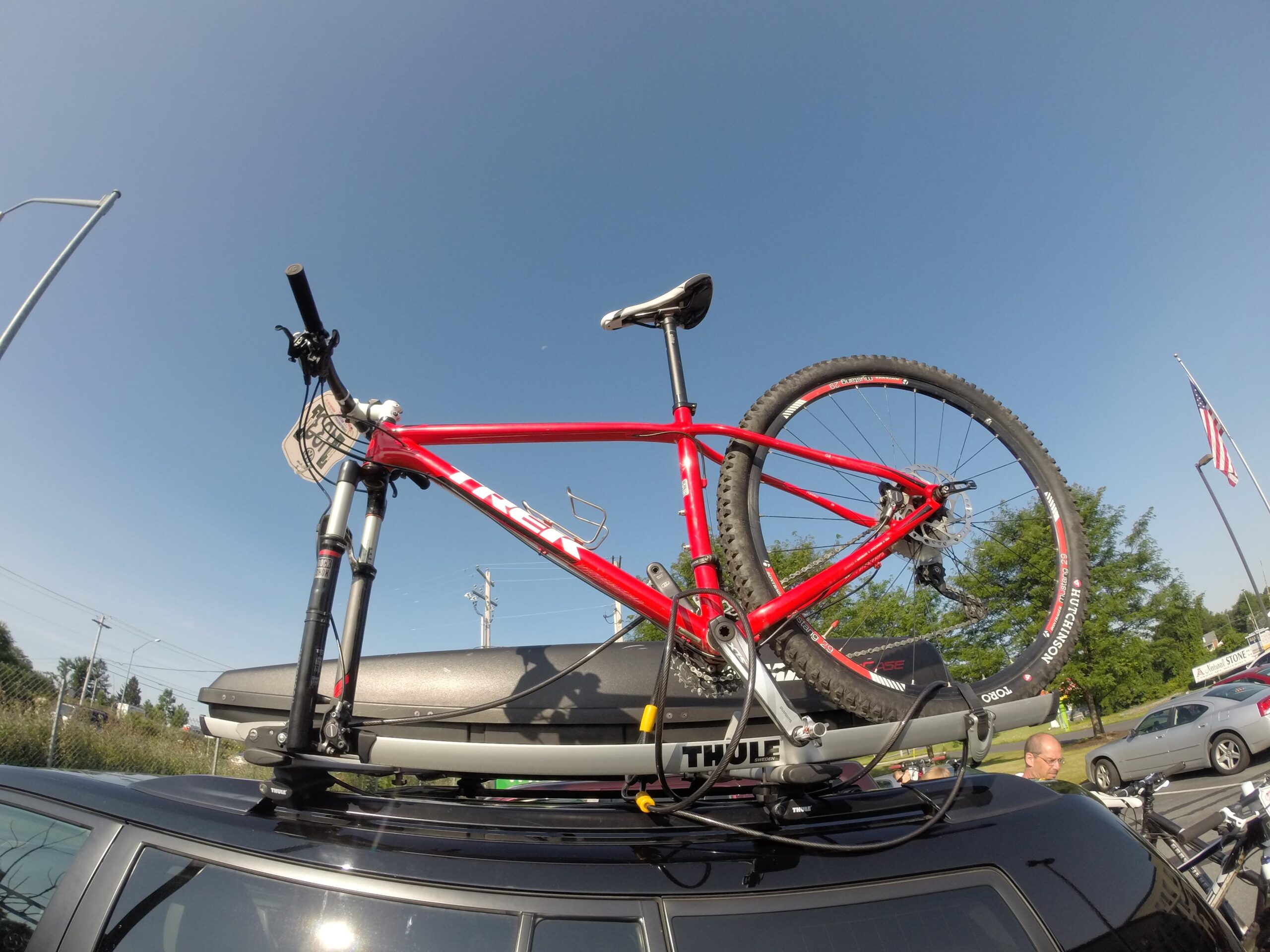 Trek Superfly 8: A red mountain bike is secured on the roof rack of a car, with a clear blue sky in the background. The bike features knobby tires and a sleek frame. A Thule bike carrier is shown mounted on the car, and several vehicles and a flag are visible in the surroundings.