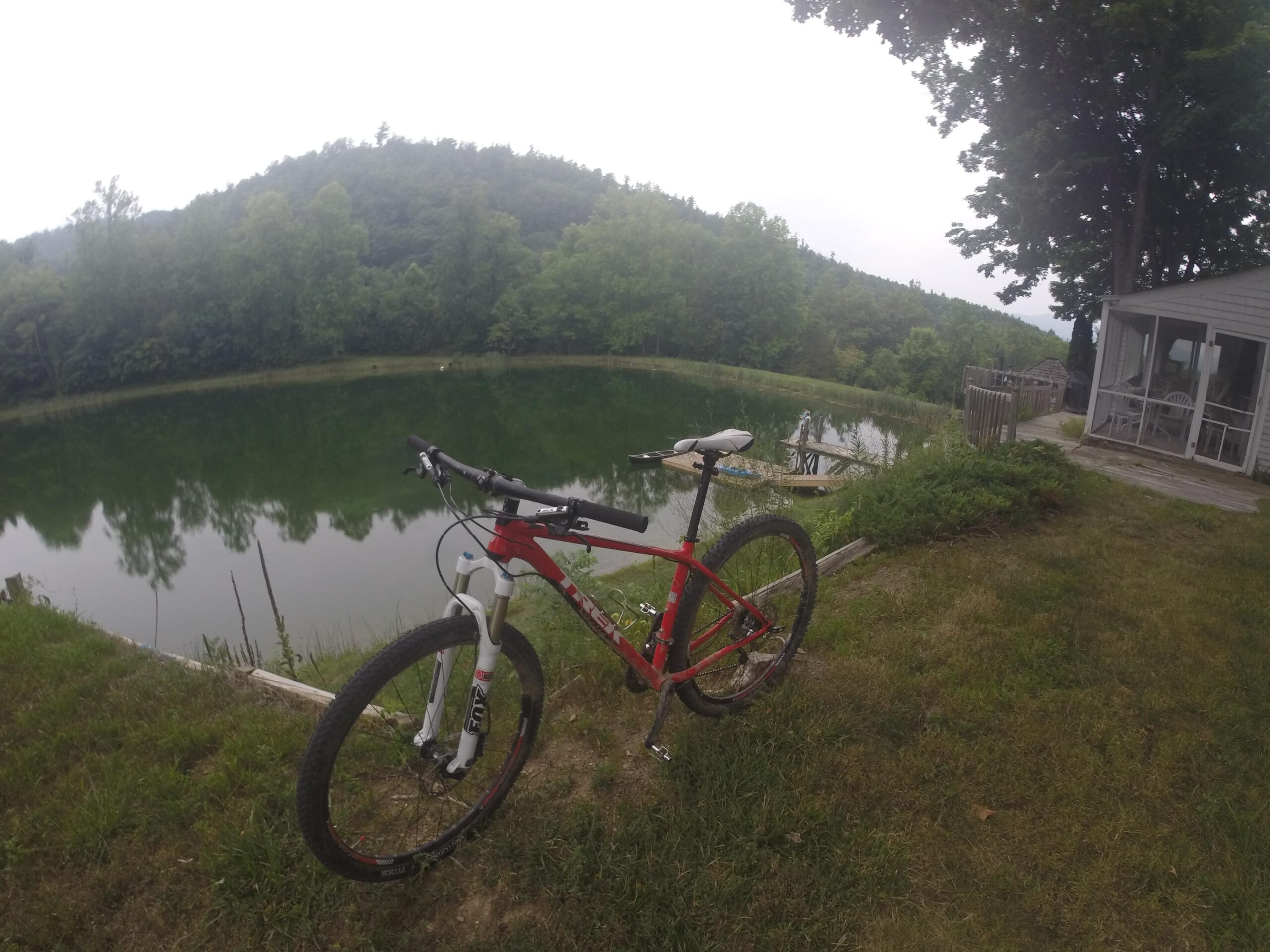 Trek Superfly 8: A red mountain bike stands on grass near a calm, reflective pond surrounded by lush greenery and trees. In the background, a small cabin can be seen, with a wooden platform by the water. The scene is slightly overcast, creating a serene and tranquil atmosphere.