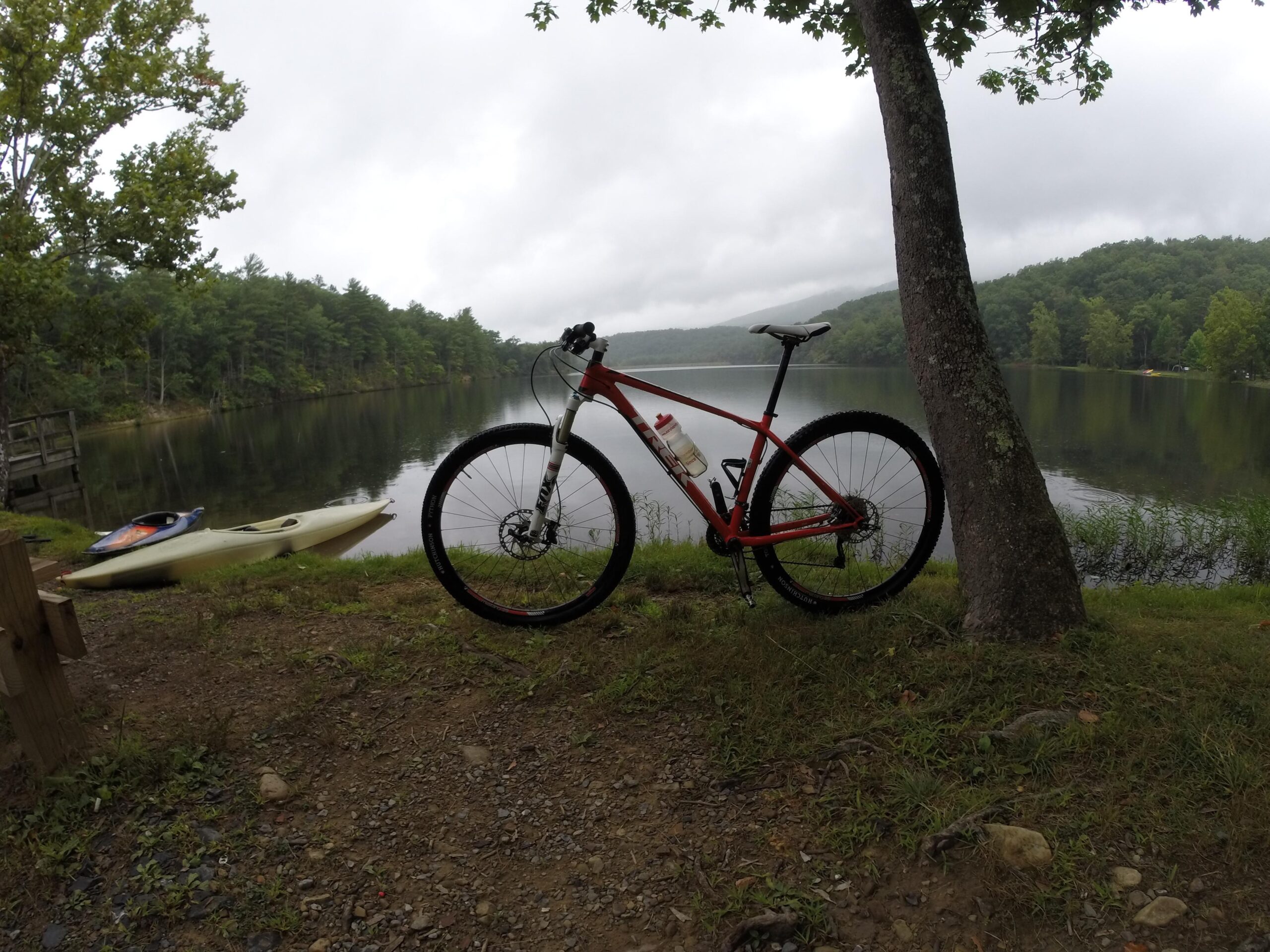 Trek Superfly 8: A red mountain bike leaning against a tree by the edge of a calm lake, with a yellow kayak in the foreground. The scene is surrounded by lush green trees, and the sky is overcast, creating a tranquil outdoor environment.