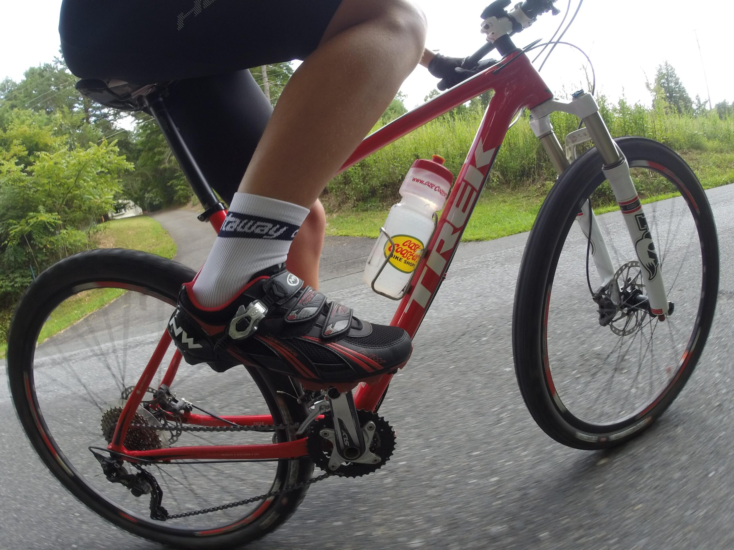 Trek Superfly 8: A close-up view of a cyclist's leg pedaling on a red Trek bicycle. The cyclist is wearing black cycling shoes and white socks with "Kentucky" printed on them. A water bottle is mounted on the bike frame. The background features a tree-lined road, suggesting an outdoor cycling environment.