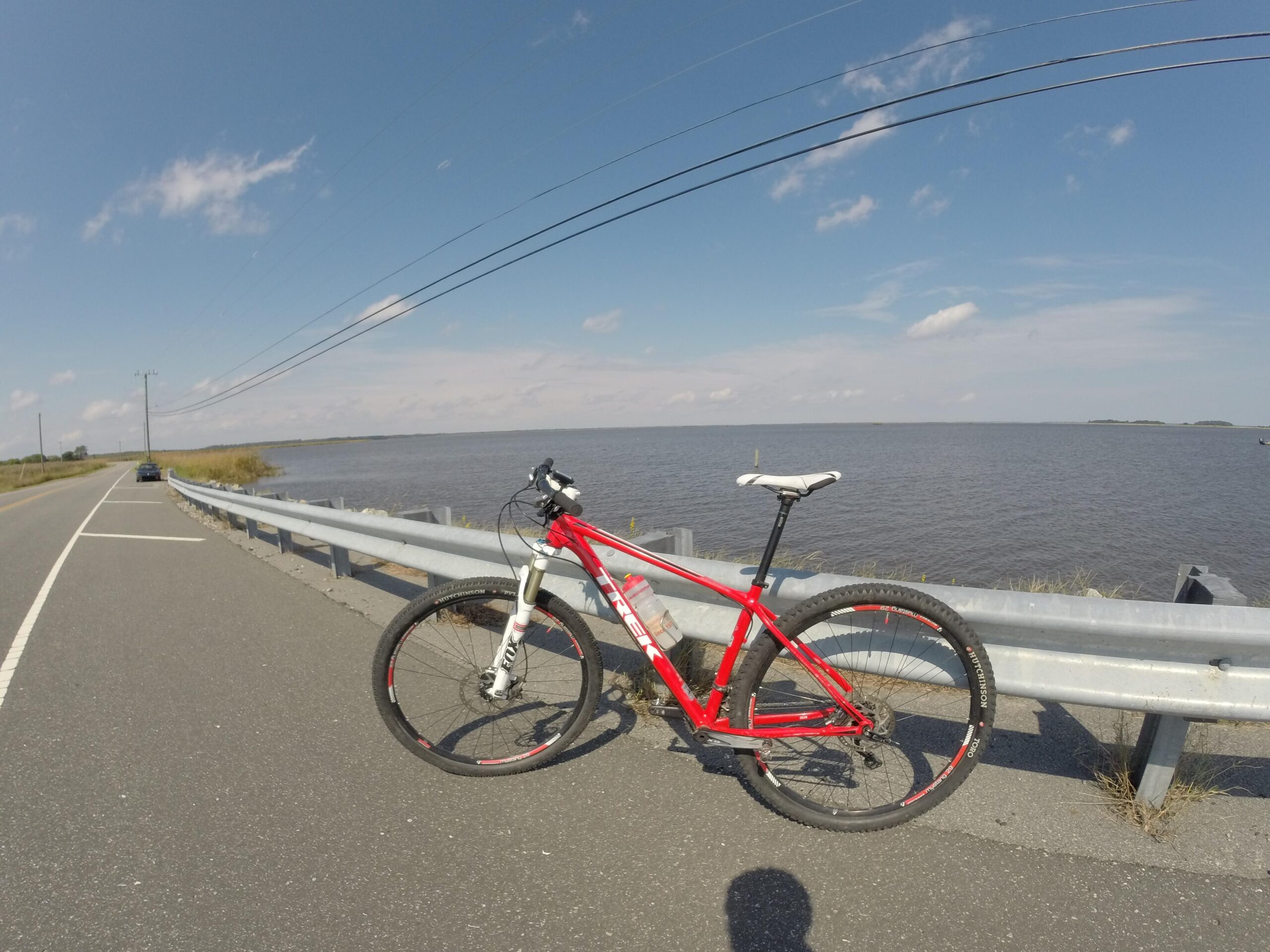 Trek Superfly 8: A red mountain bike parked beside a guardrail along a road, with a view of a body of water under a clear blue sky. In the background, there is a single car on the road and grassy areas lining the water's edge.