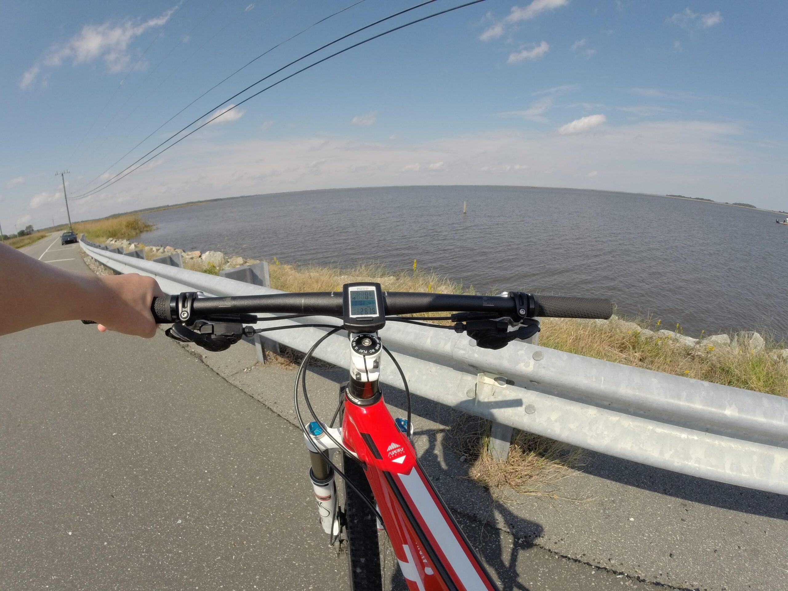 Trek Superfly 8: A view from the handlebars of a red mountain bike, with a hand gripping the handlebars and a digital display visible. In the background, a calm body of water stretches to the horizon under a bright blue sky with scattered clouds. A paved road runs alongside the water, bordered by a metal guardrail, with grass and rocks visible nearby.