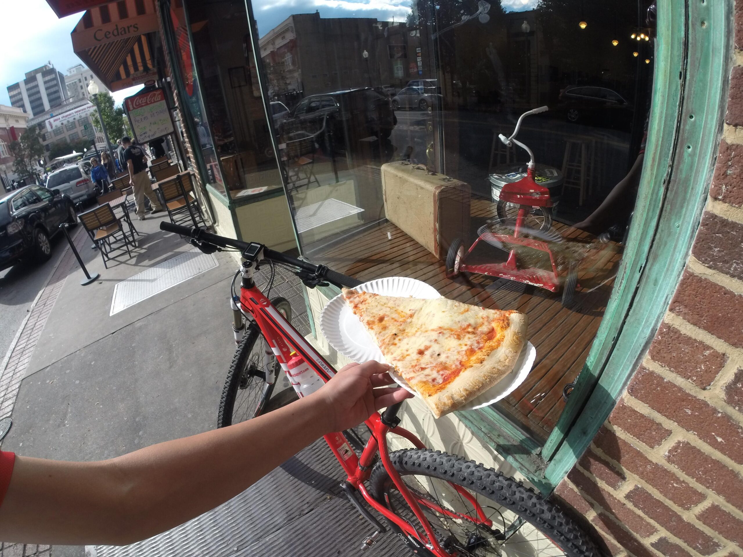 Trek Superfly 8: A hand holding a slice of pizza on a paper plate, positioned next to a red bicycle parked on the sidewalk. The photo captures a storefront with large windows, revealing a glimpse of the interior. In the background, people stroll by on the street, and outdoor seating is visible. A vintage red toy car is reflected in the window.