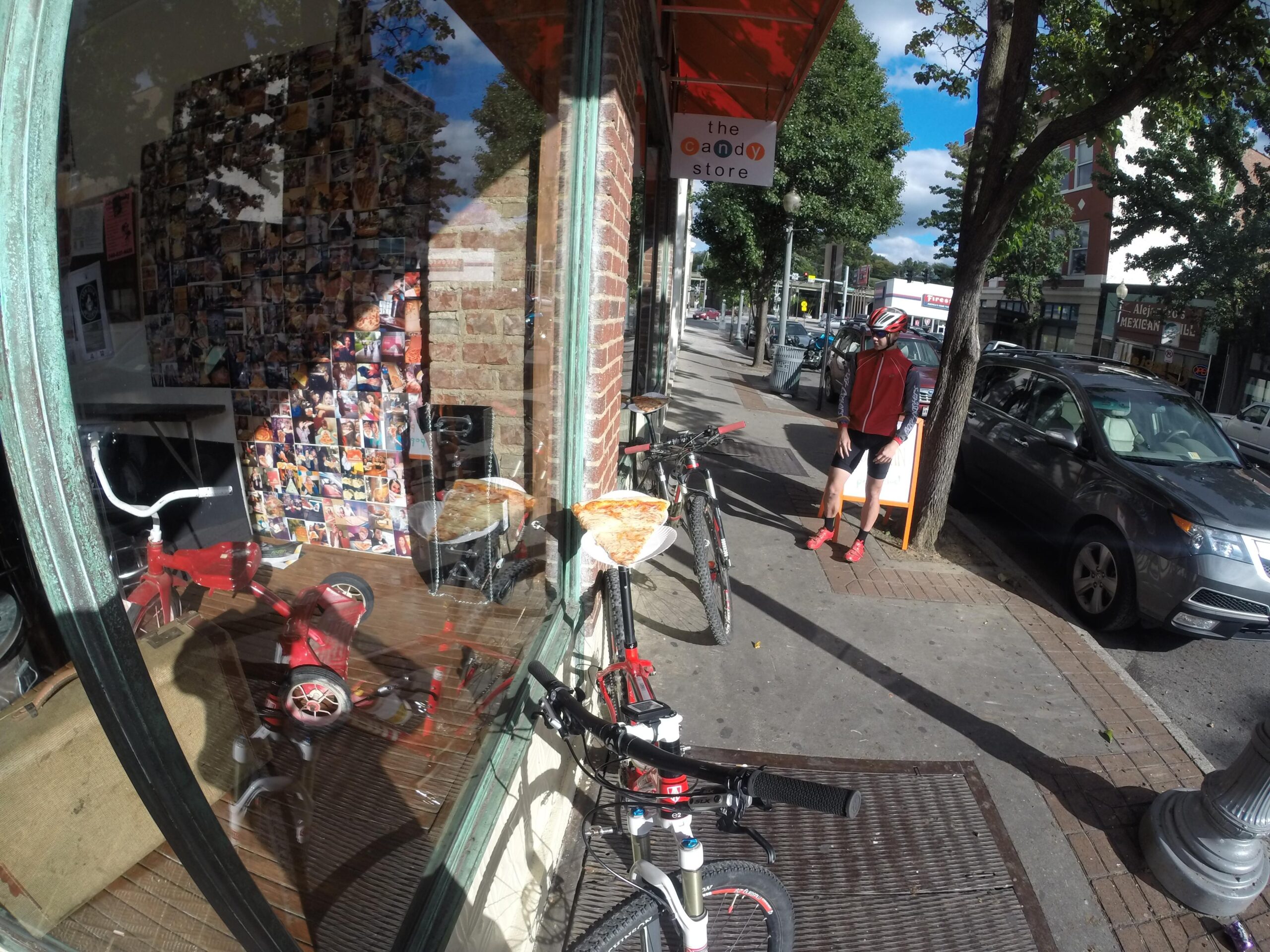 Trek Superfly 8: A view through the window of a store showcasing a wall adorned with various photographs. Outside, a small red tricycle and two bicycles are parked on the sidewalk. A person in cycling attire stands nearby, with an orange store awning visible above. Trees and a city street fill the background, adding to the lively urban atmosphere.