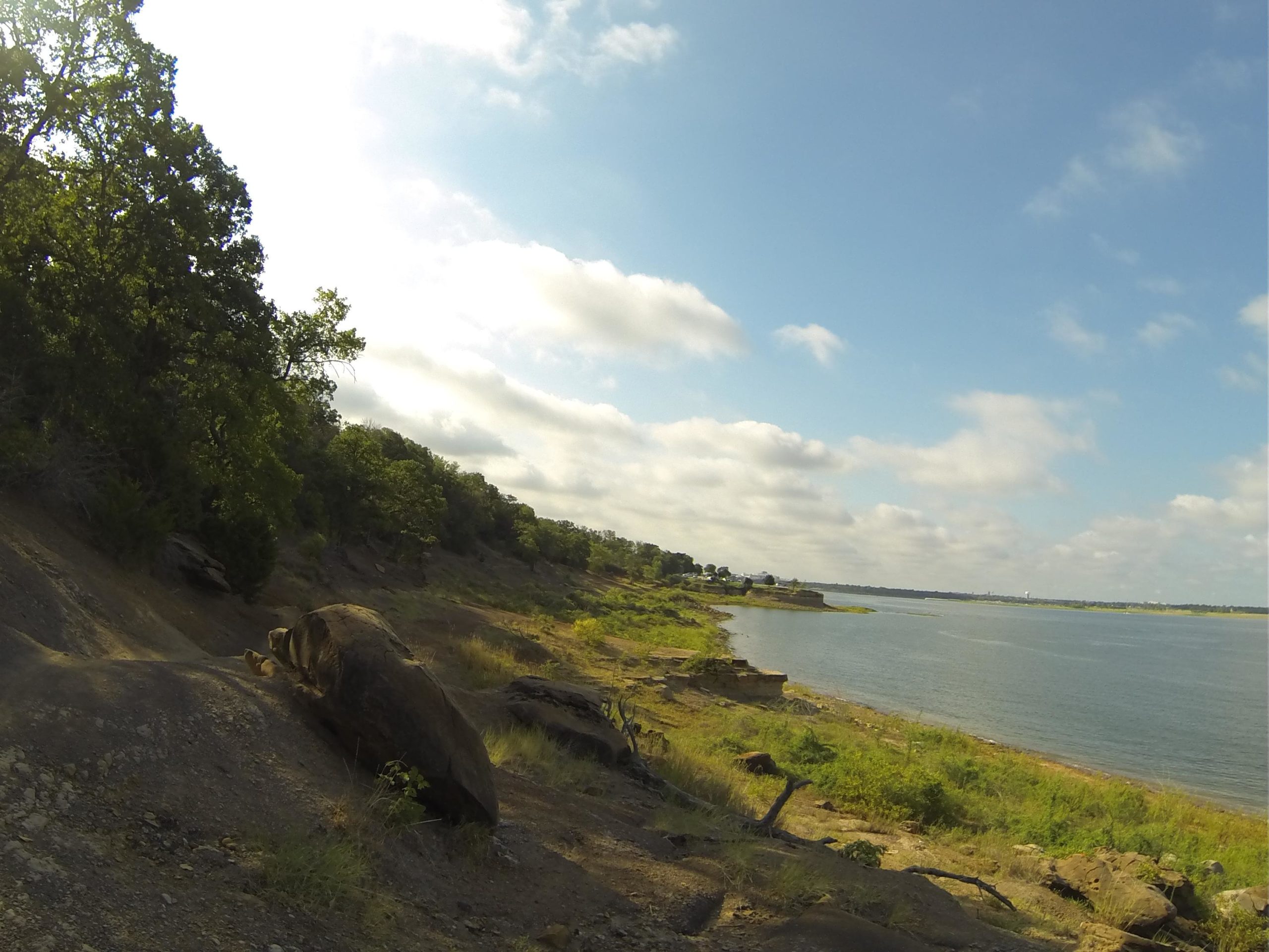 A serene landscape featuring a rocky shoreline along a calm lake, with green trees and grassy areas in the foreground. The sky is partly cloudy, creating a peaceful outdoor scene. Northshore Trail mountain bike trail.