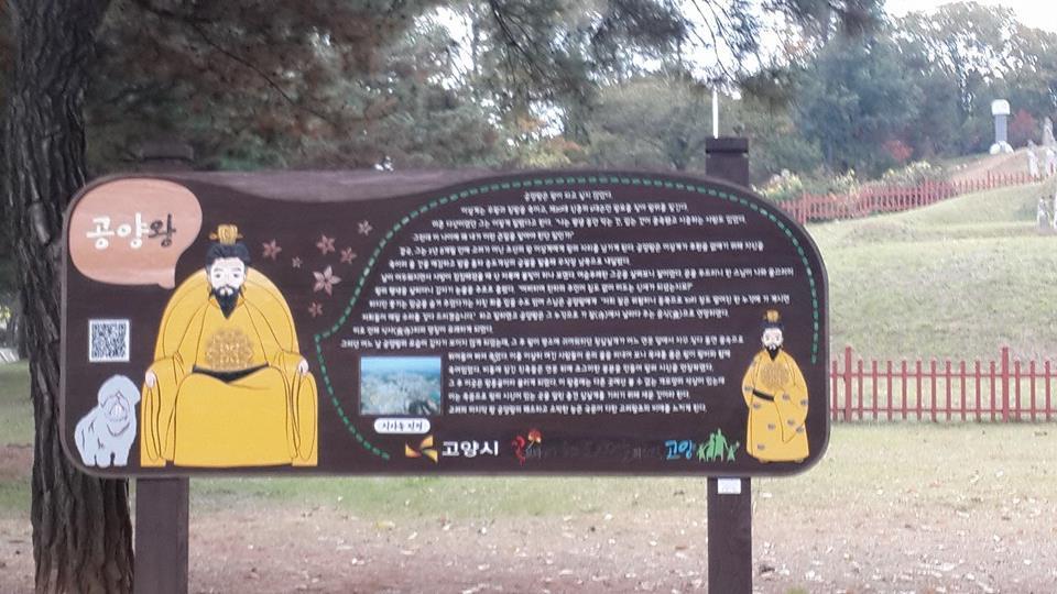 A wooden signboard featuring an illustrated depiction of a historical figure sitting on a throne, accompanied by a stylized drawing of a dog. The sign contains written text in Korean, likely providing information about the figure and the location. The background shows a grassy area with a fence and trees. Myoji mountain bike trail.