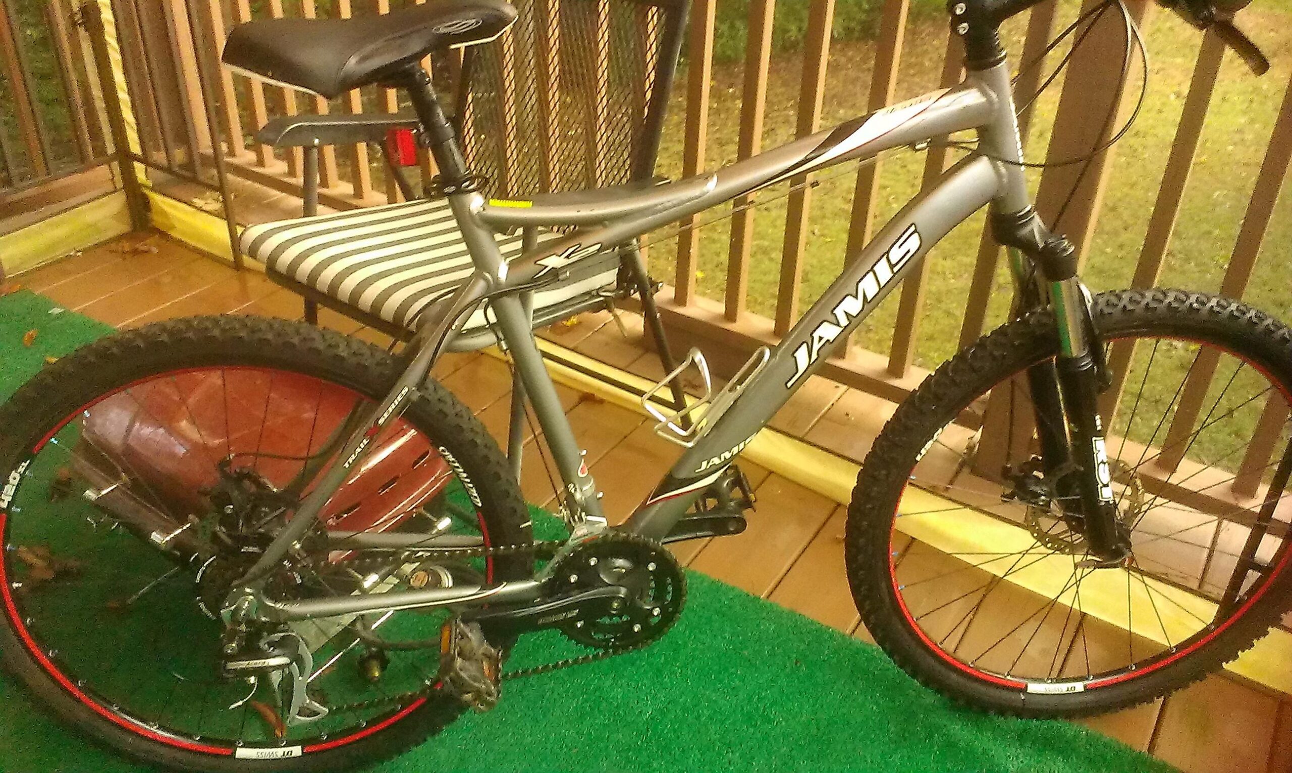 Jamis X2: A silver Jamis mountain bike with red accents is parked on a porch with a green rug. In the background, there's a striped chair and a grassy area visible through the railing.