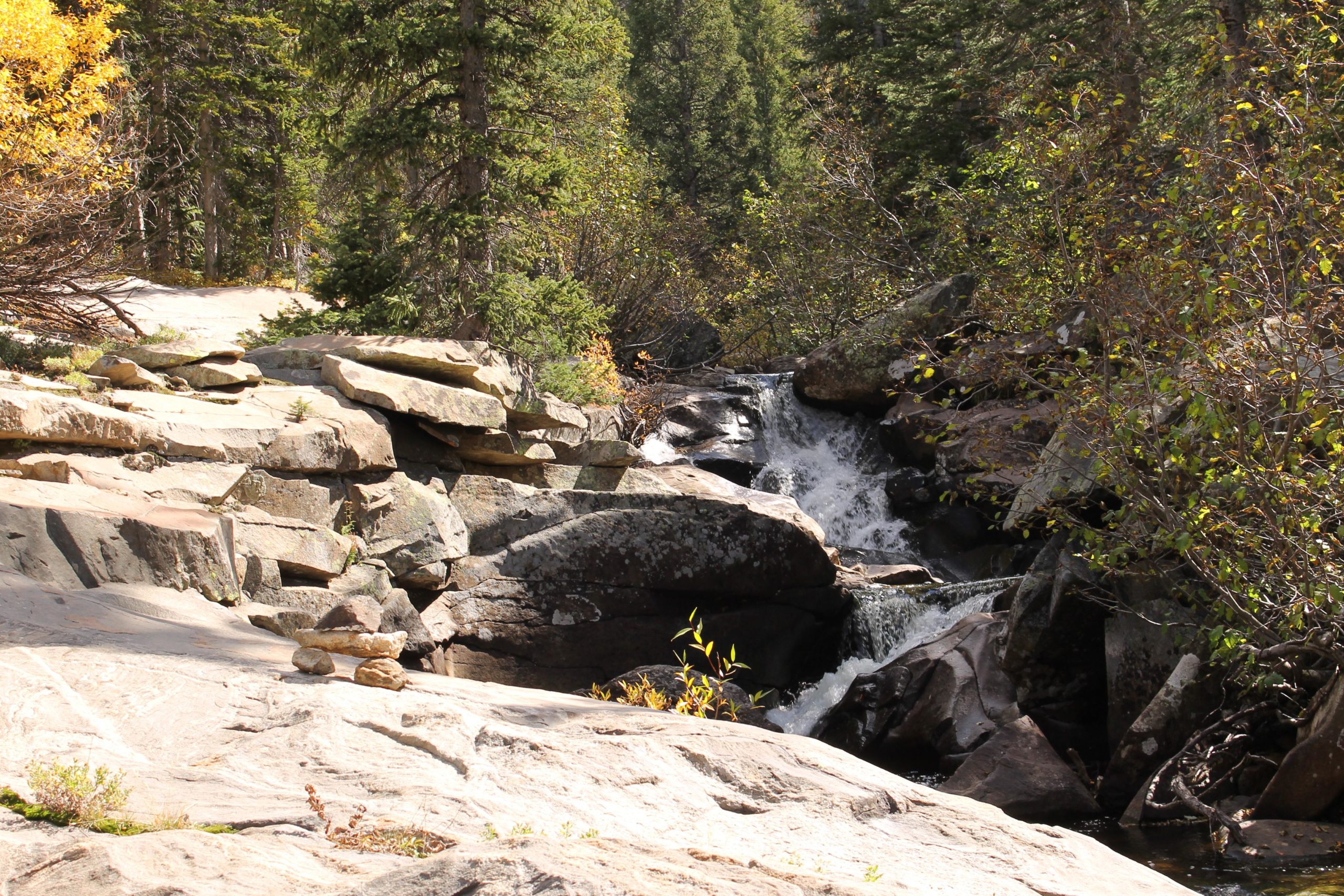 A rocky landscape featuring a small waterfall cascading over boulders, surrounded by green trees and autumn foliage. Sunlight filters through the foliage, highlighting the textures of the stones and the flowing water. Fish Creek Falls mountain bike trail.
