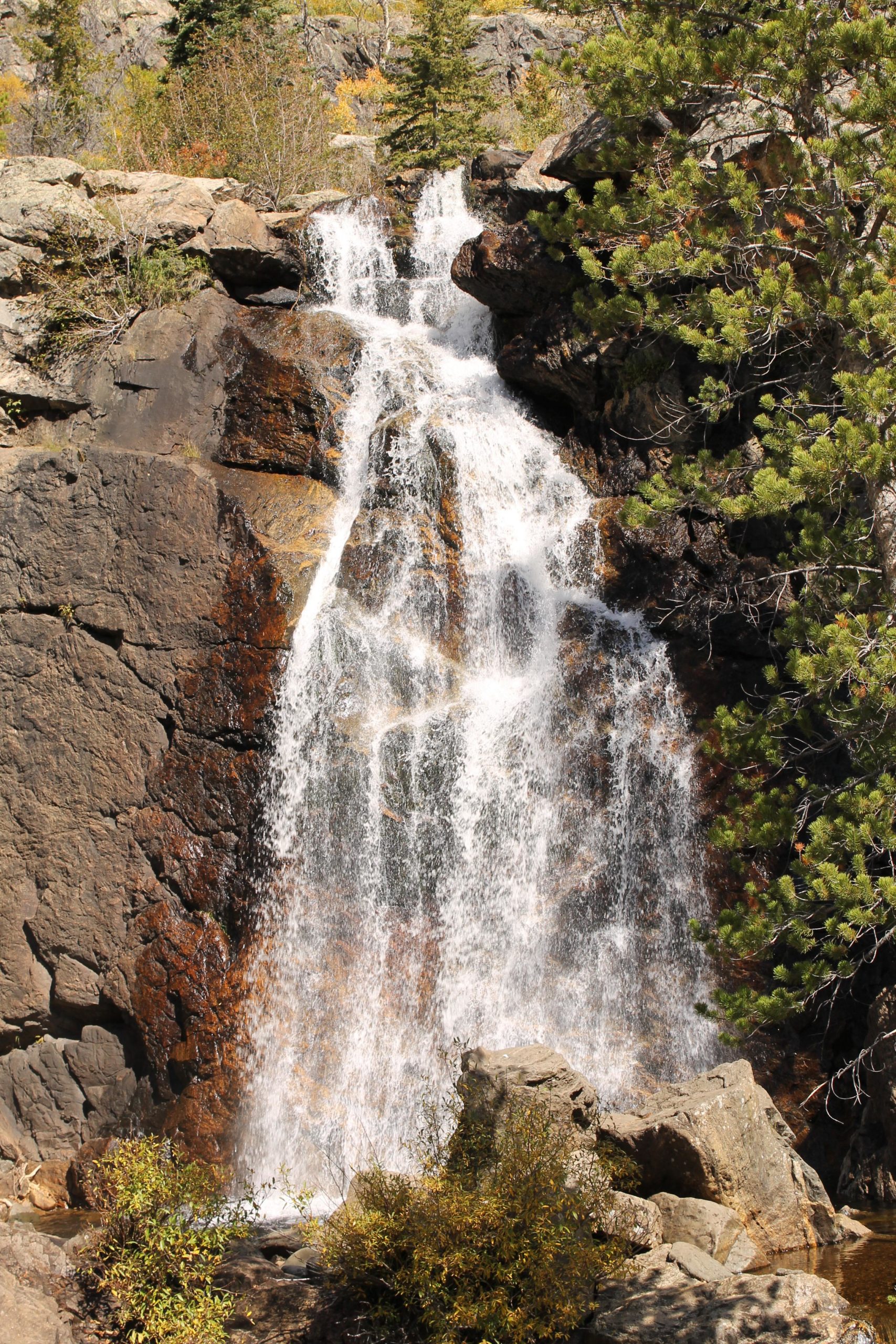 A cascading waterfall flowing down a rocky cliff, surrounded by greenery and autumn foliage. Water splashes over the rocks, creating a mist at the base. Fish Creek Falls mountain bike trail.
