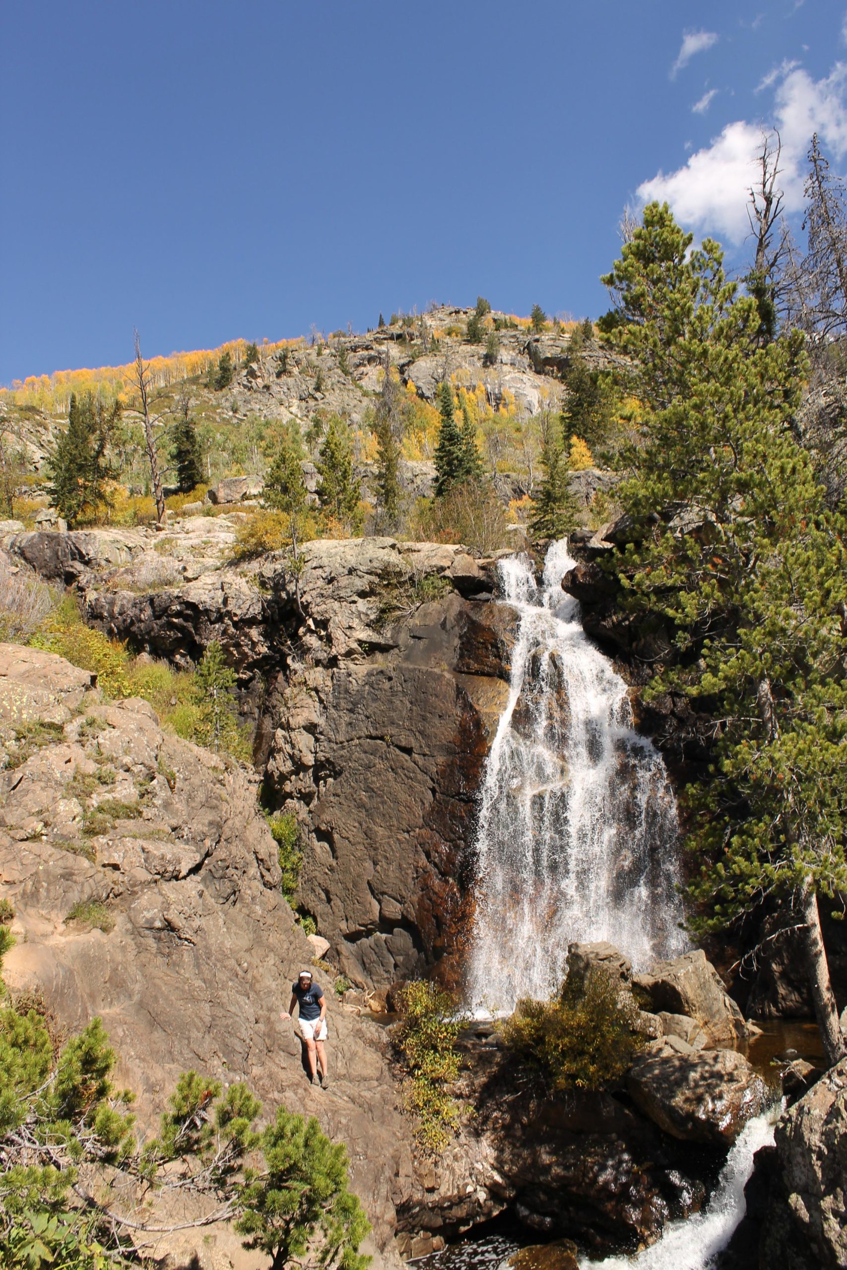 A scenic view of a waterfall cascading down rocky cliffs, surrounded by lush greenery and trees. A person is hiking along the rocky terrain, with a clear blue sky above and colorful autumn foliage visible on the mountainside in the background. Fish Creek Falls mountain bike trail.