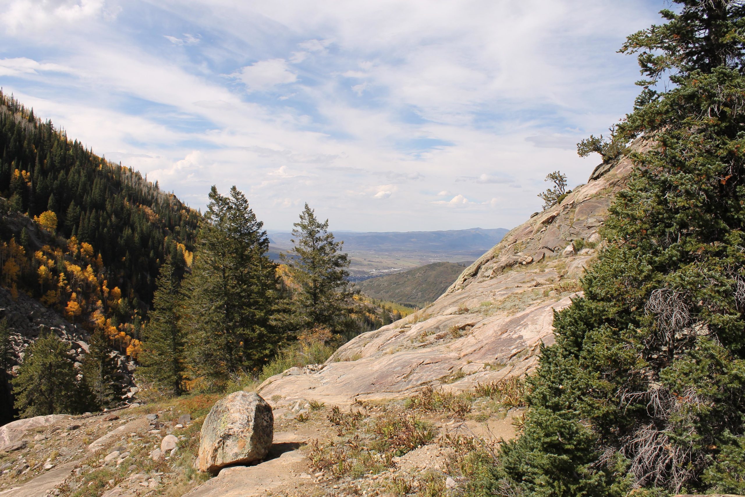 A scenic view of a mountainous landscape featuring rocky terrain and evergreen trees, with vibrant autumn foliage in the valley below. The sky is partly cloudy, showcasing a blend of blue and white. Fish Creek Falls mountain bike trail.