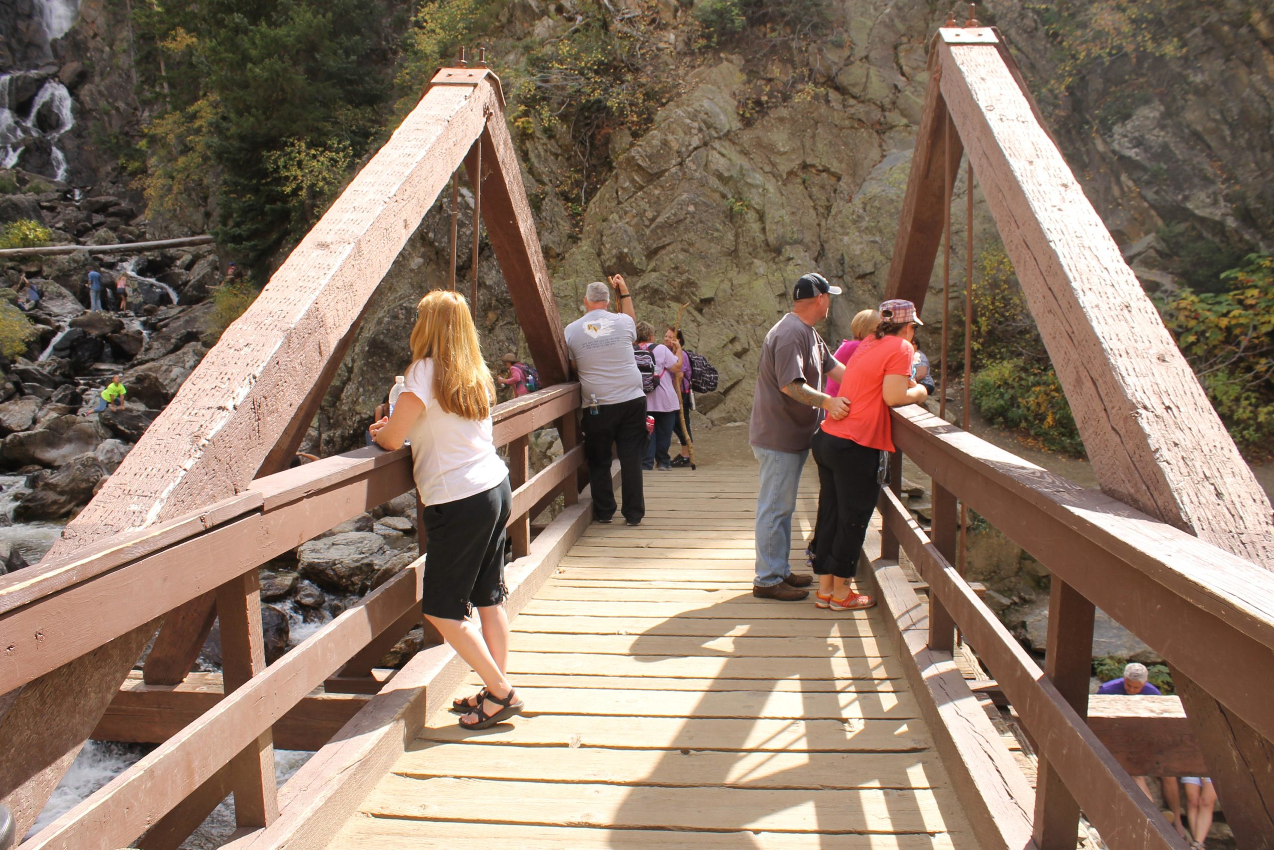 A wooden bridge crosses a rocky landscape, with several people standing on it, looking at the scenery below. Some appear to be enjoying the view, while others are engaged in conversation. In the background, a waterfall cascades down the rocks, surrounded by greenery. The scene captures a peaceful outdoor moment in nature. Fish Creek Falls mountain bike trail.