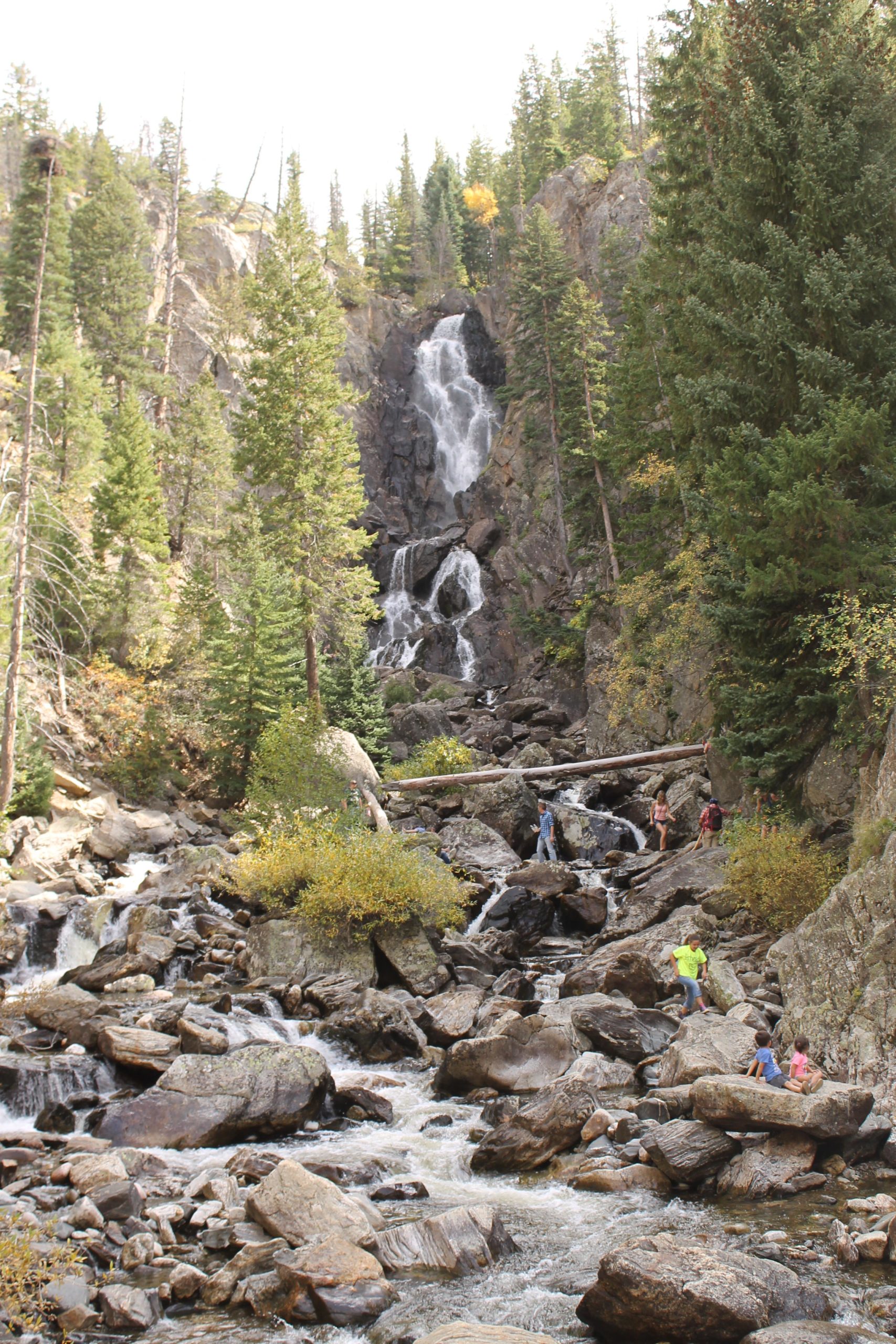 A scenic view of a waterfall cascading down rocky terrain surrounded by lush green pine trees. Several people are visible near the water's edge, exploring the area and relaxing on the rocks. Vibrant autumn foliage can be seen among the greenery. Fish Creek Falls mountain bike trail.