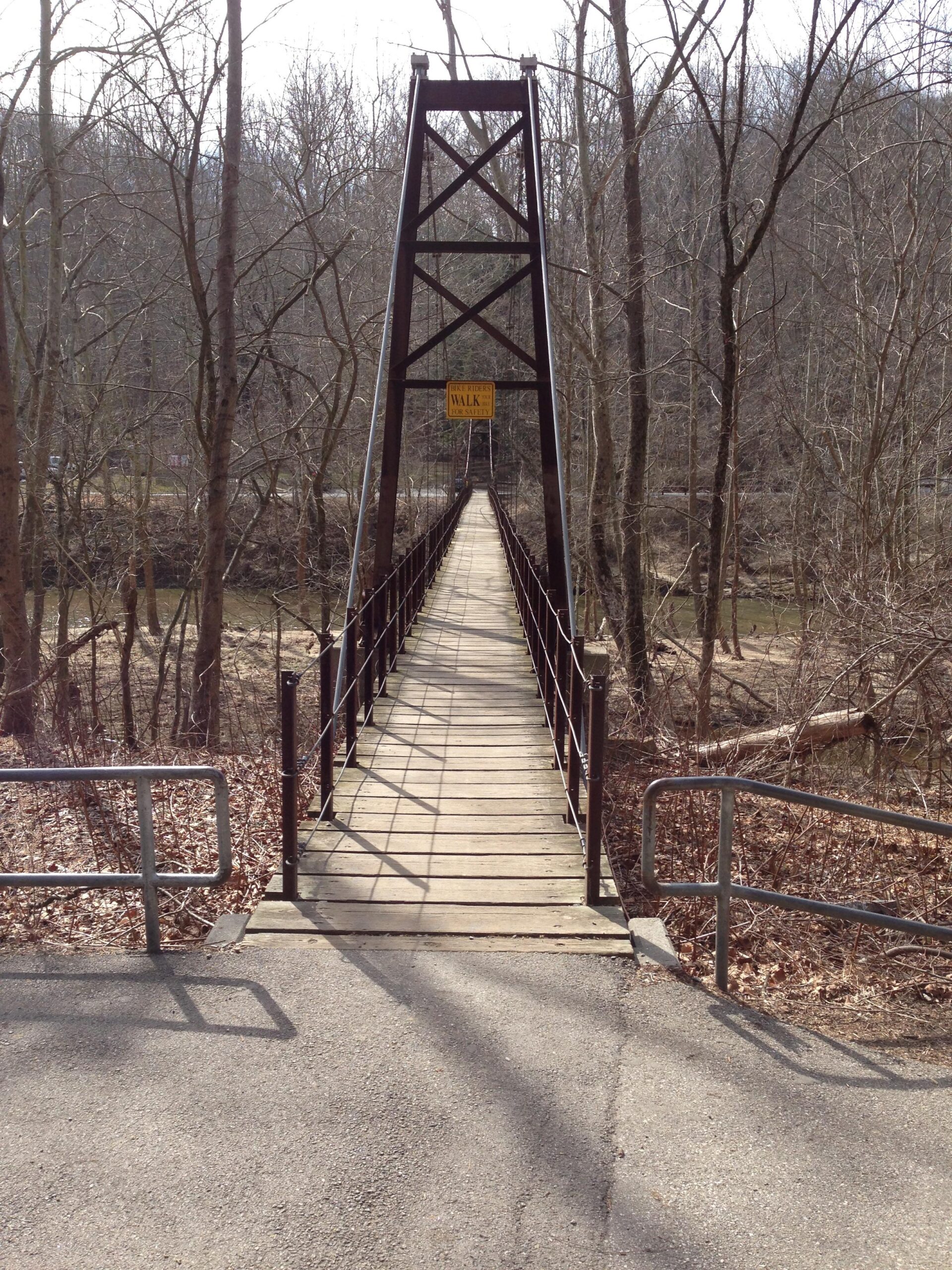 Alt text: A suspension bridge made of wood and metal spans over a narrow river, surrounded by bare trees in an early spring landscape. A yellow "WALK" sign is visible at the entrance of the bridge, with a gravel path leading up to it. The ground is covered with fallen leaves and the atmosphere is calm and quiet. Patapsco Valley State Park (Avalon Area) mountain bike trail.