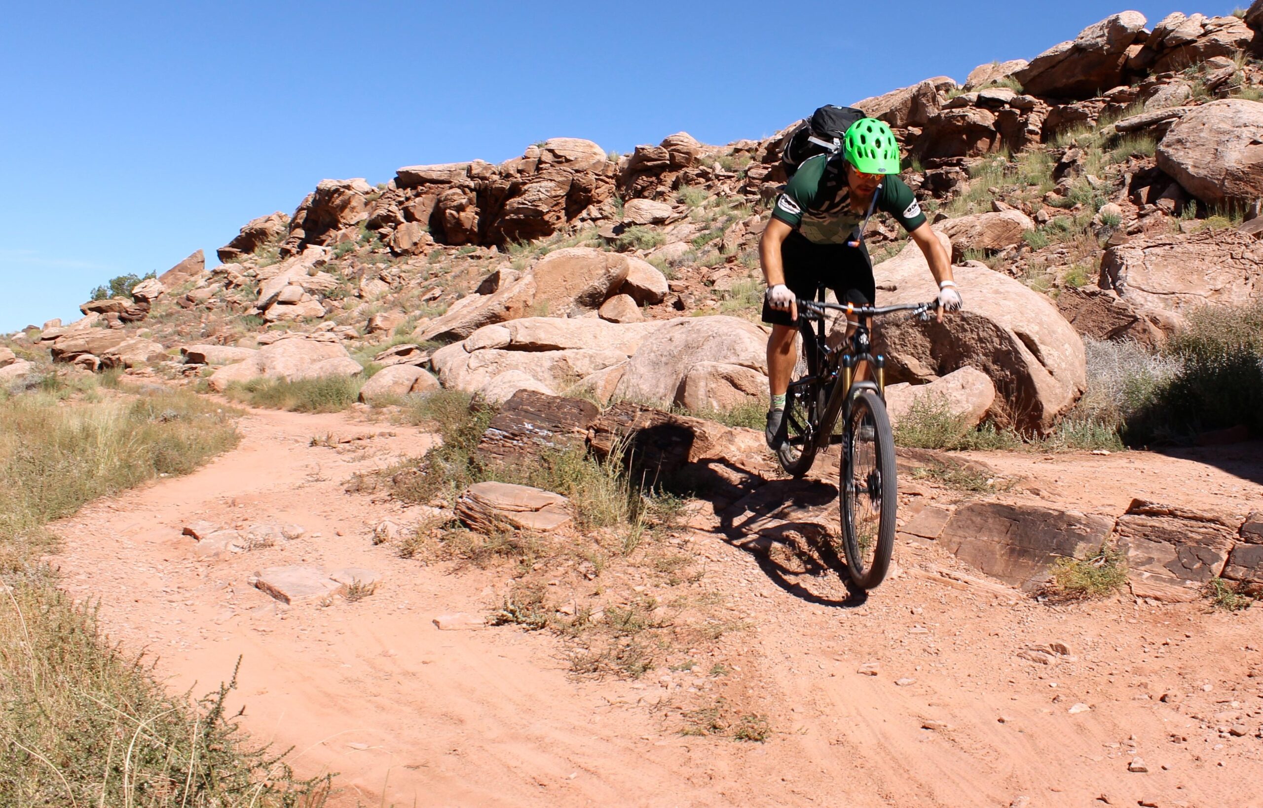 A mountain biker navigates a rugged dirt trail surrounded by large rocks and shrubs under a clear blue sky. The cyclist wears a green helmet and a green and black outfit, showcasing an adventurous ride in a natural terrain. Moab Brand Trails mountain bike trail.