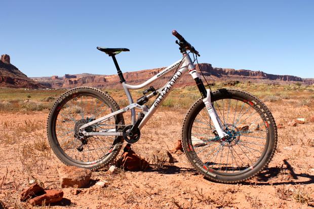 Durango Bike Company Moonshine 650b: A mountain bike standing on rocky terrain with a scenic landscape of cliffs and mesas in the background under a clear blue sky.
