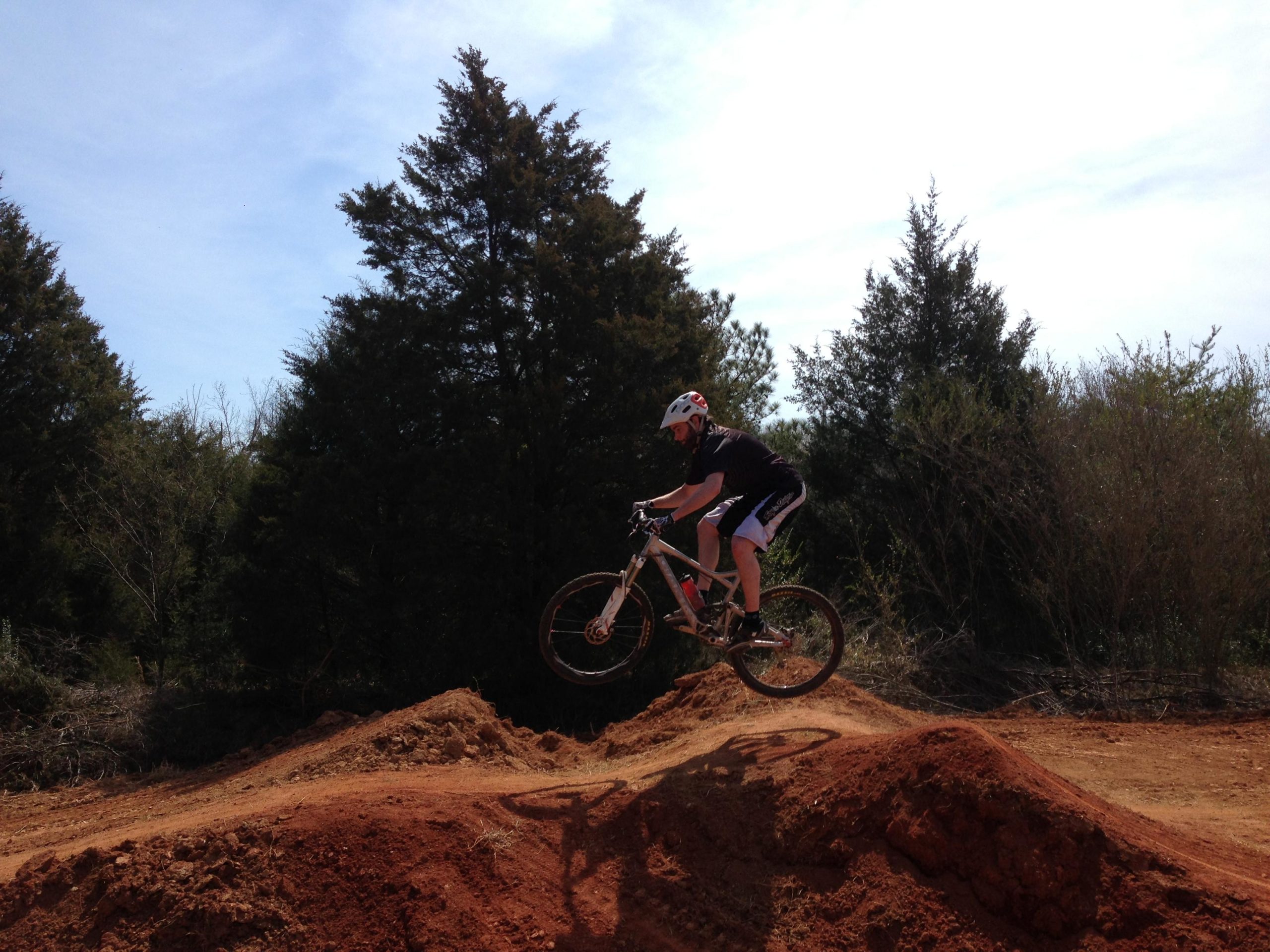 A person in a helmet and athletic attire is mid-air while performing a jump on a mountain bike over a dirt ramp, surrounded by trees and a clear blue sky. Signal Hill mountain bike trail.