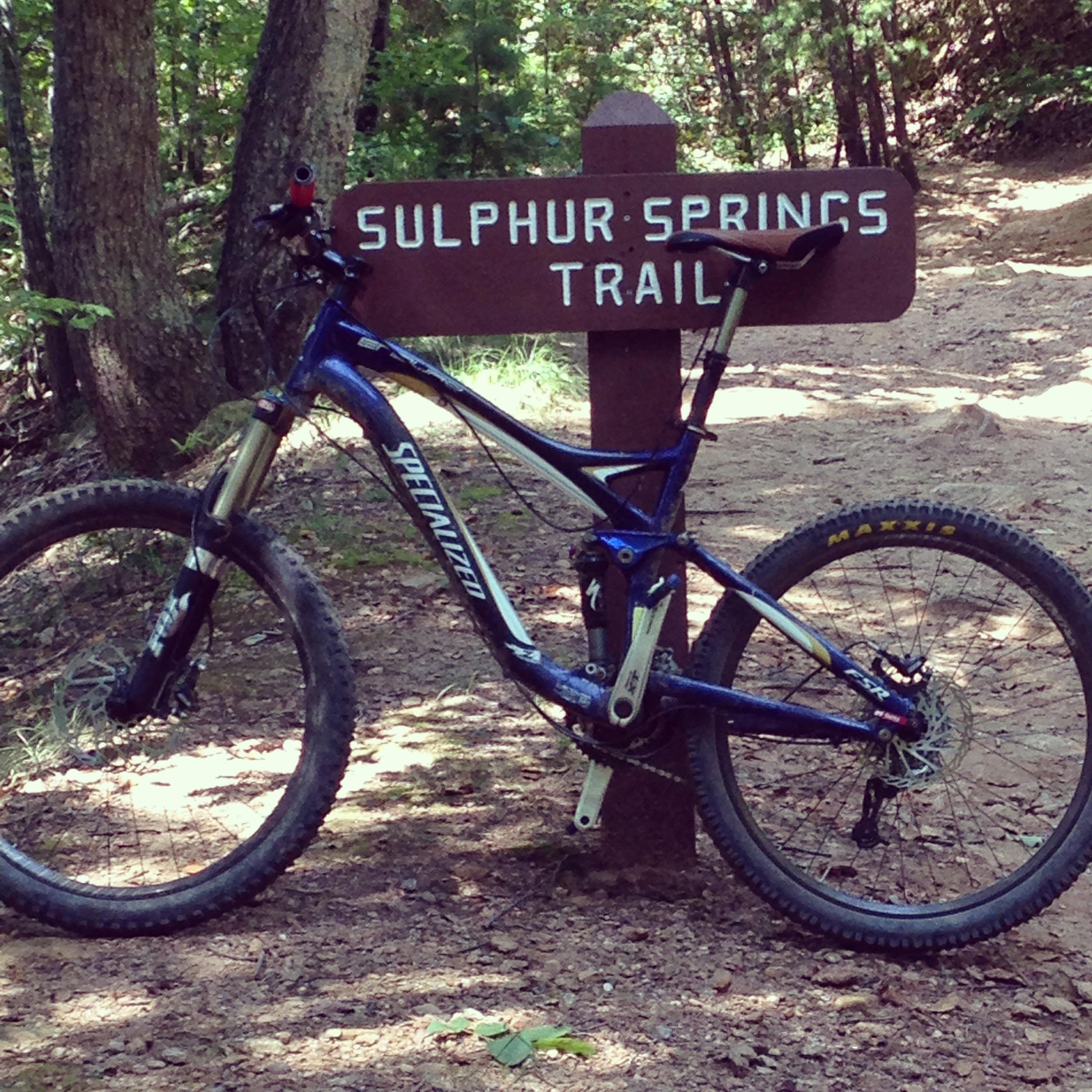 Specialized Enduro SL Pro: A blue mountain bike leaning against a wooden sign that reads "Sulphur Springs Trail", surrounded by trees in a dirt path setting.