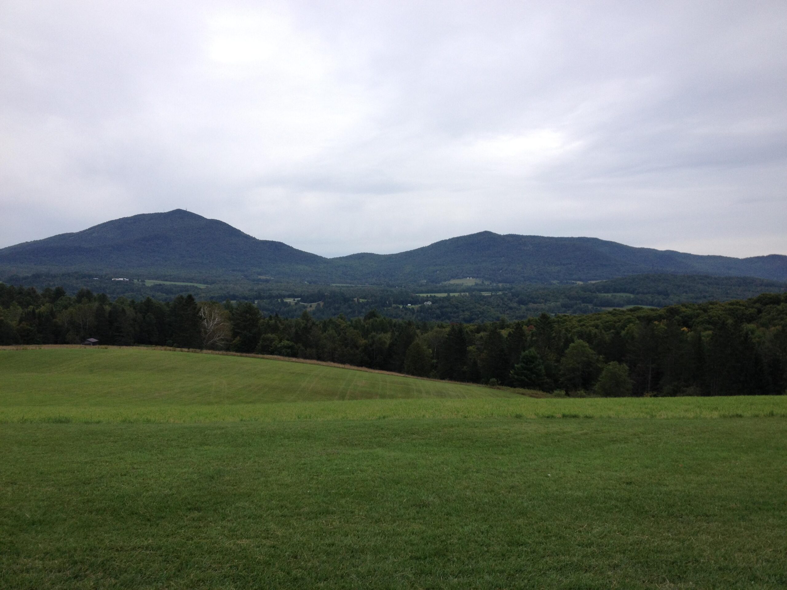 A tranquil landscape featuring rolling green hills in the foreground, leading to two prominent mountains in the background under a cloudy sky. The scene captures a lush, wooded area with diverse vegetation, creating a serene natural setting. Kingdom Trails mountain bike trail.