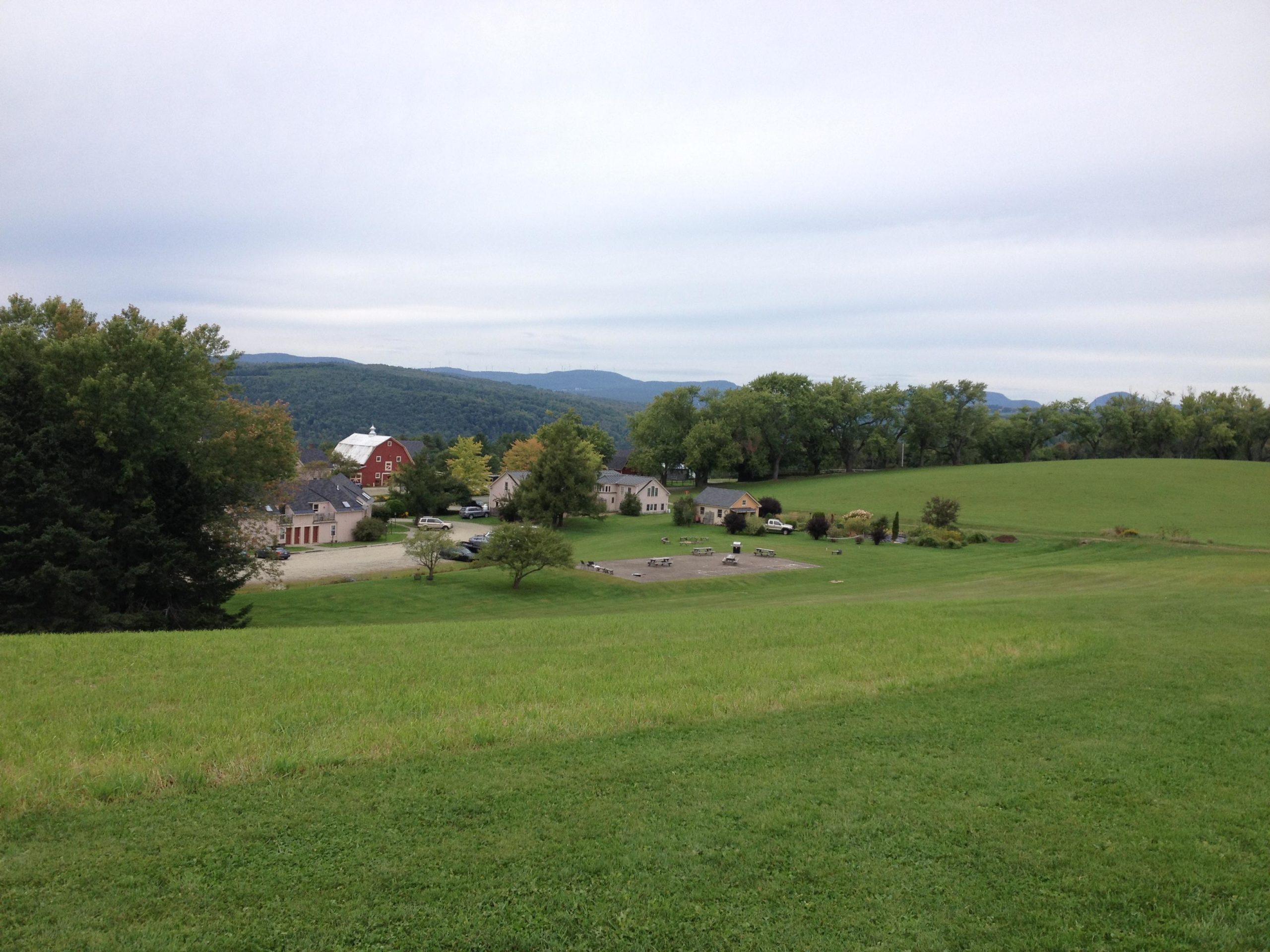 A picturesque landscape showing a serene, green field leading down to a small cluster of houses and a barn, with rolling hills in the background under a cloudy sky. Kingdom Trails mountain bike trail.