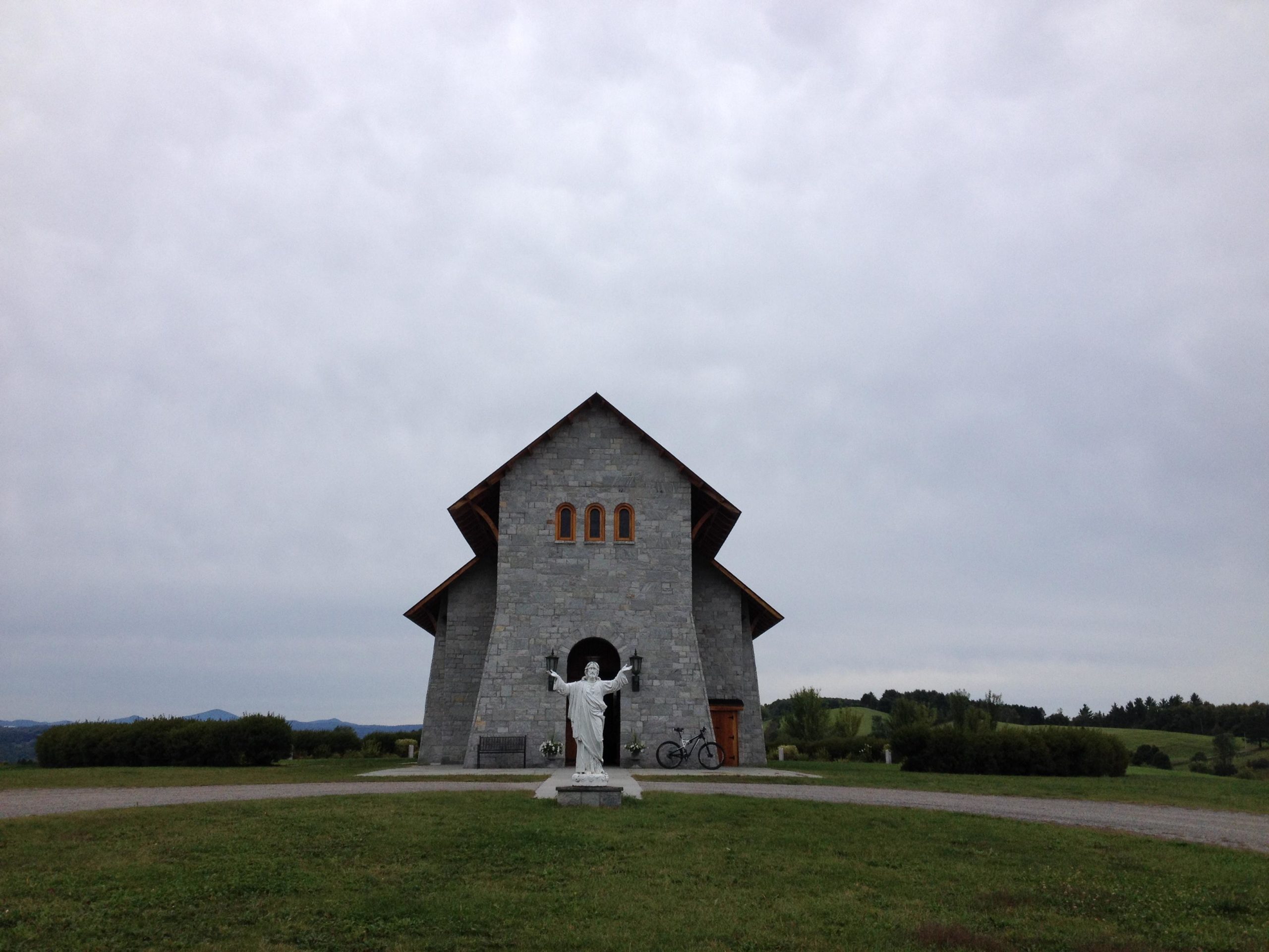 A gray stone chapel with wooden accents stands against a cloudy sky. In front of the chapel, there is a white statue of a figure extending its arms. A bicycle is parked next to the building, surrounded by green grass and distant rolling hills. Kingdom Trails mountain bike trail.